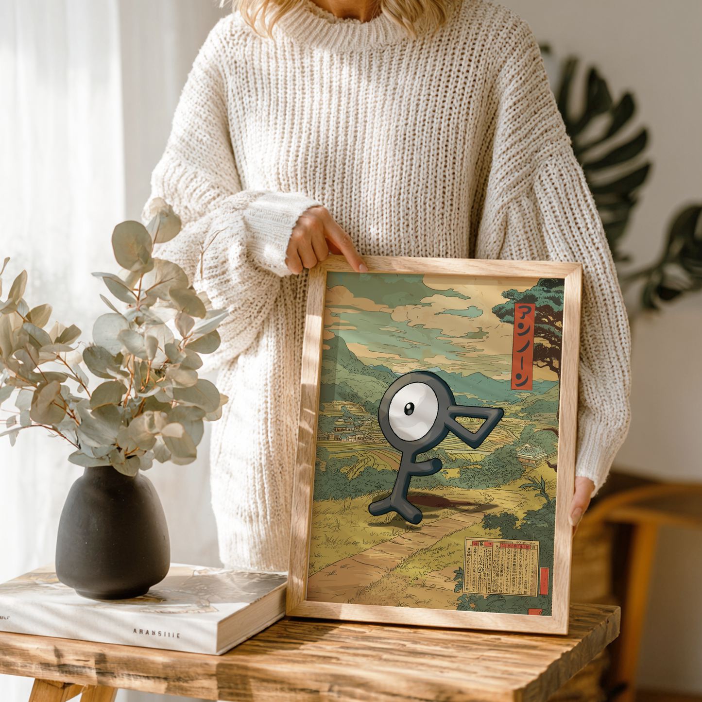 Person holding a framed artwork with a stylized Unown Pokemon character on a wooden table.