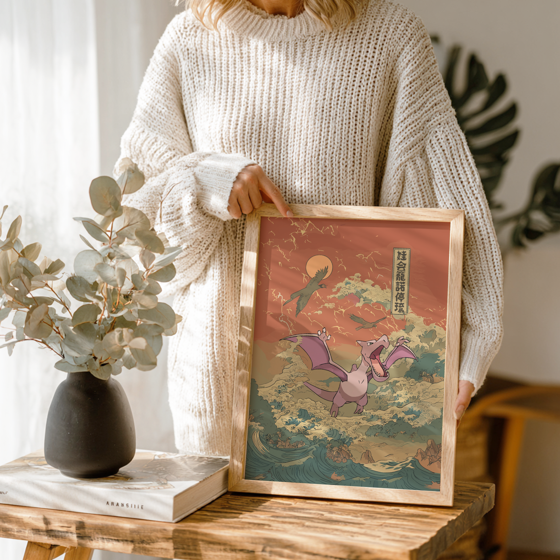 Person holding a framed illustration of Aerodactyl Pokemon on a wooden table with a vase and book in the background.