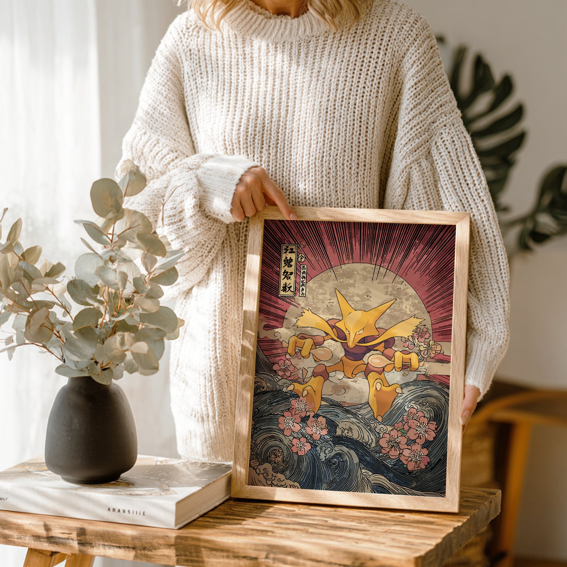 Person holding a framed artwork of a stylized Alakazam Pokemon in a room with plants and decor.