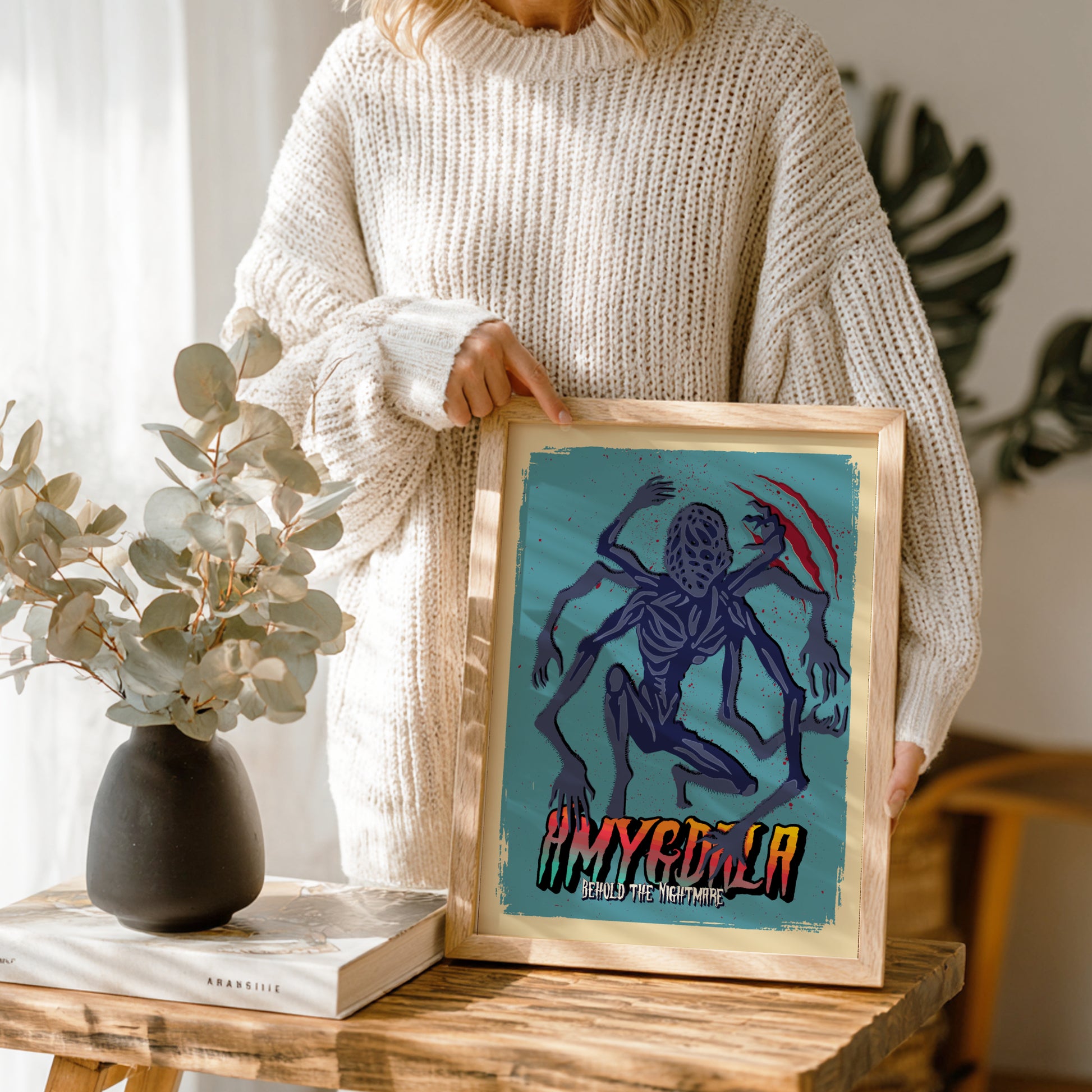 Person holding a framed artwork of Amygdala from Bloodborne Game with a blue background and text, standing in a room with a vase and plant.