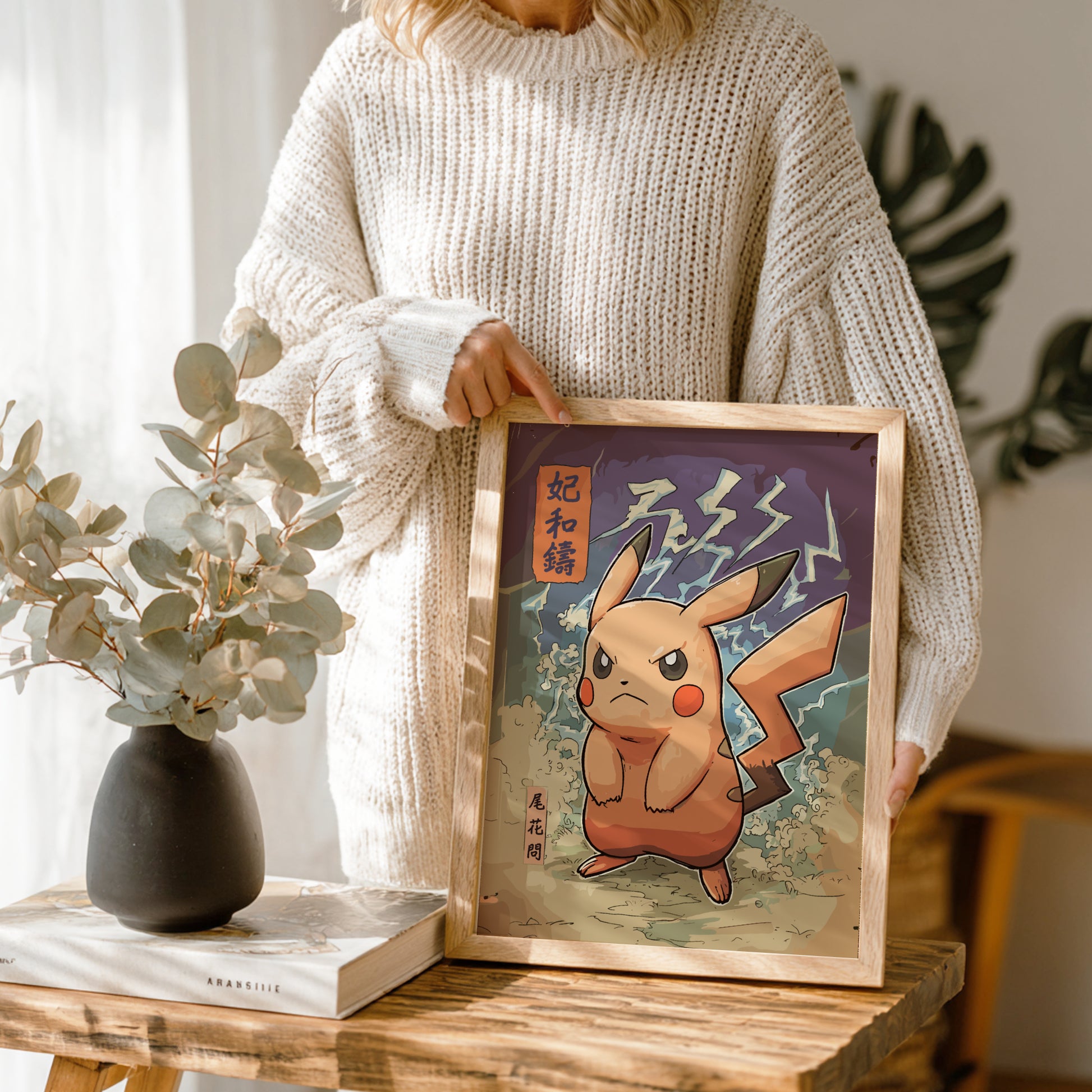 Person holding a framed artwork of an Angry Pikachu Pokemon on a wooden table with a vase and book in the background.