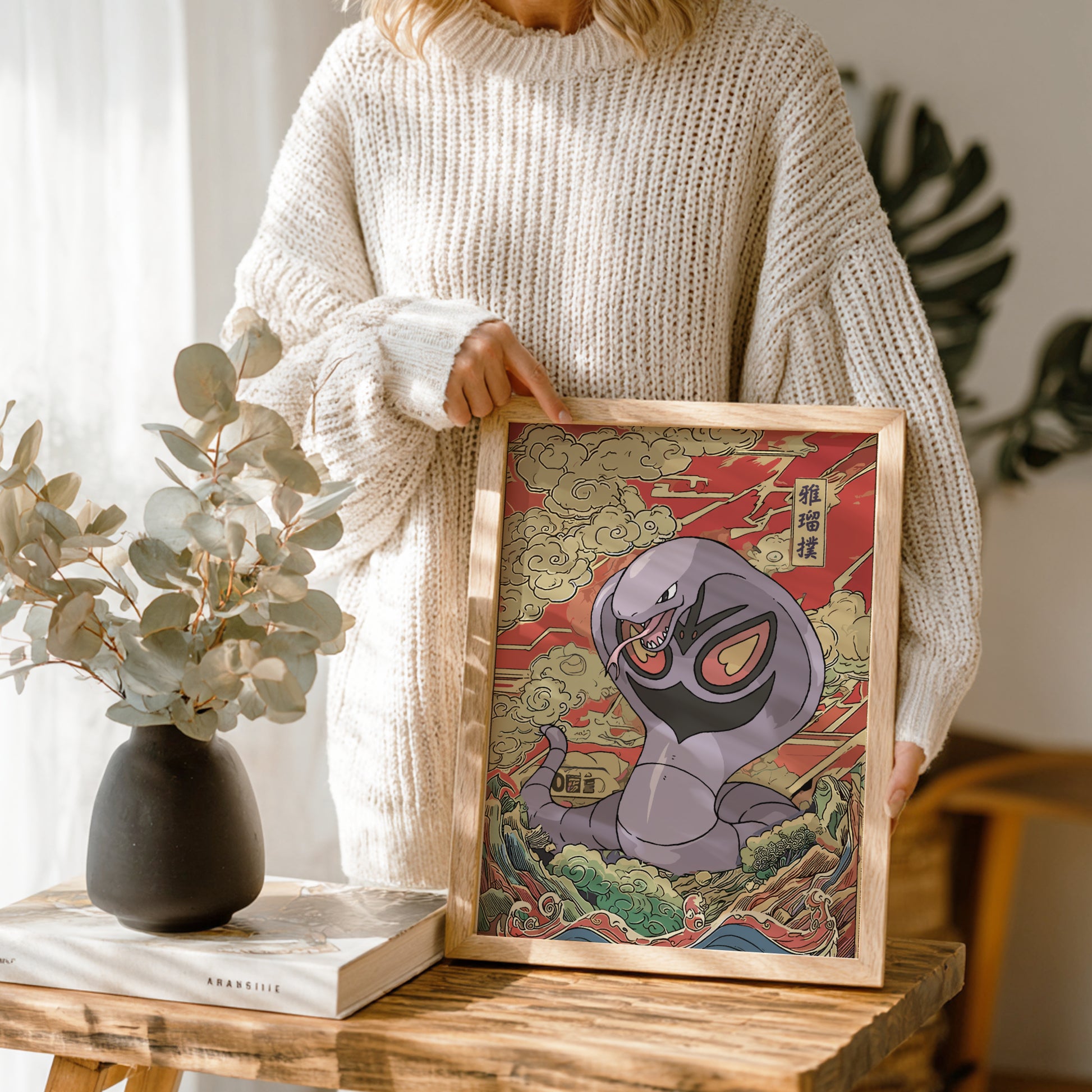 Person holding a framed artwork of Arbok Pokemon on a wooden table with a vase and book in the background.