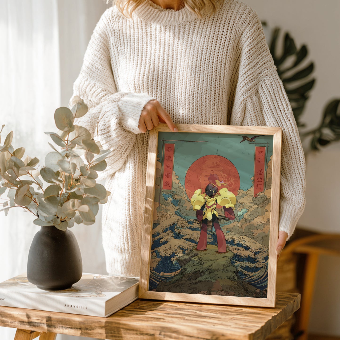 Person holding a framed artwork on a wooden table with a vase and books in the background.