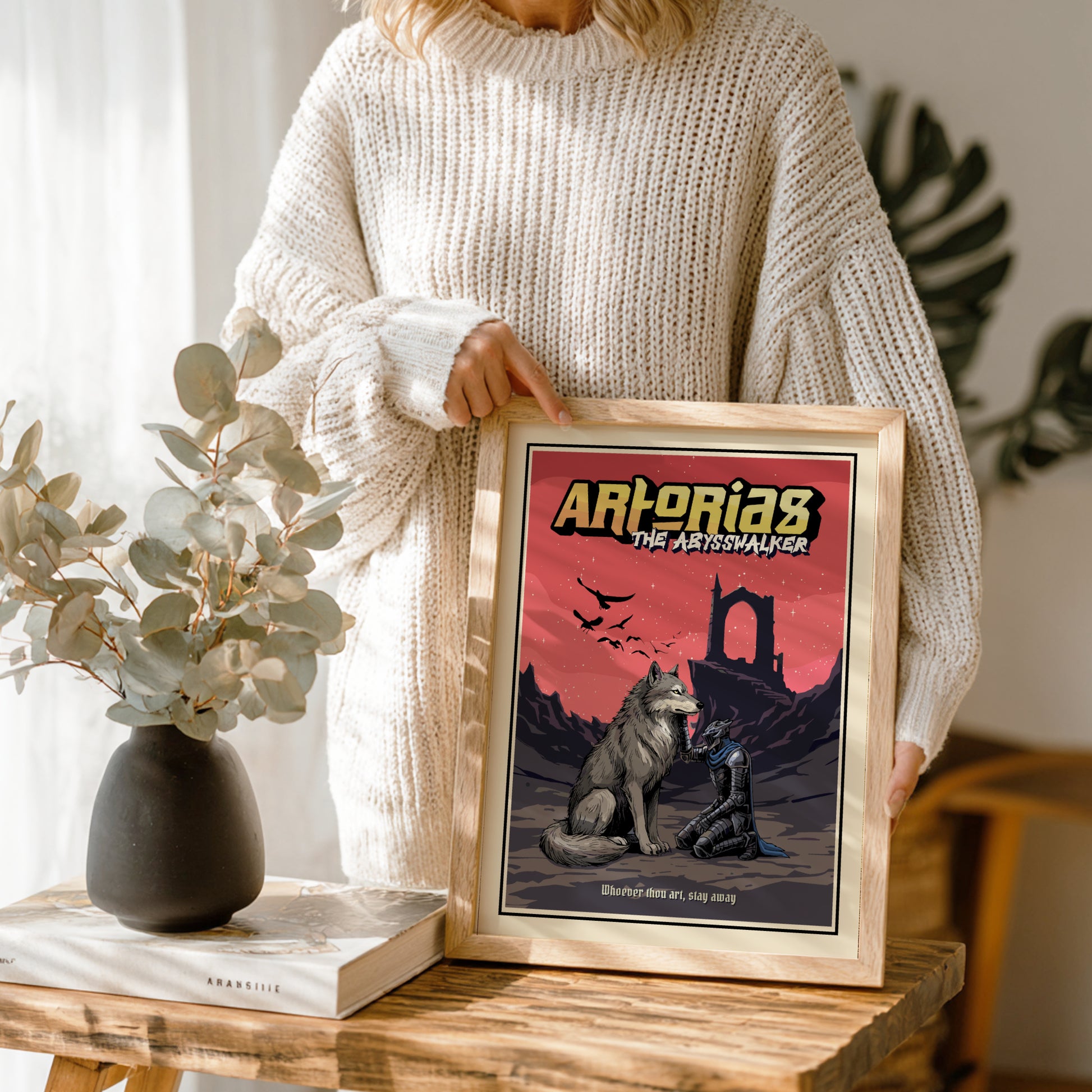 Person holding a framed poster of Artorias from Dark Souls on a wooden table with decor items.