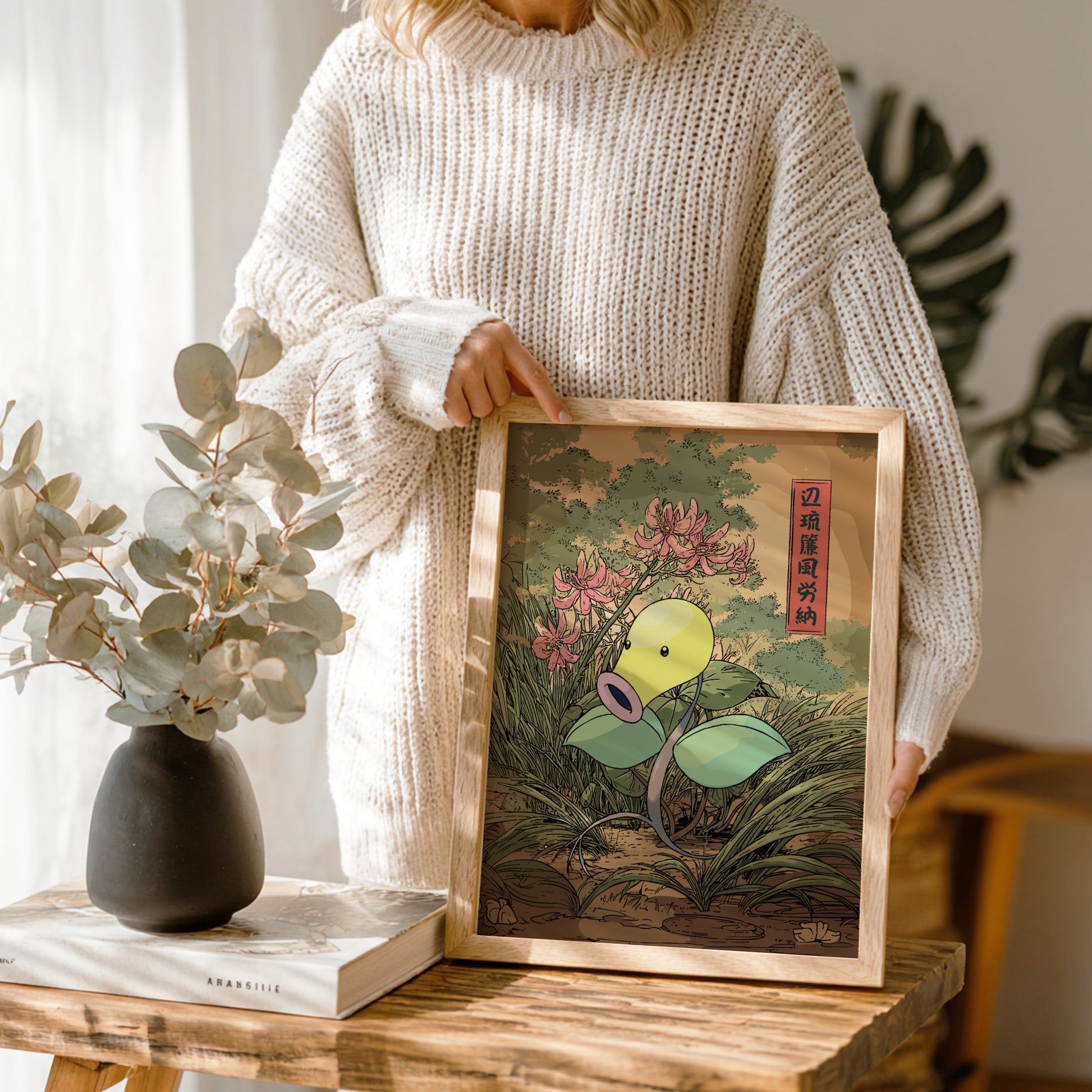 Person holding a framed artwork of Bellsprout Pokemon with flowers in a room setting.