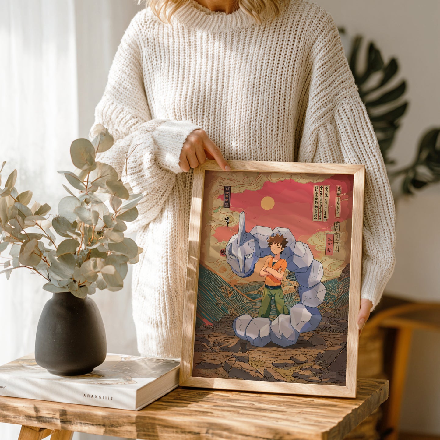 Person holding a framed artwork on a wooden table with a vase and book in the background.