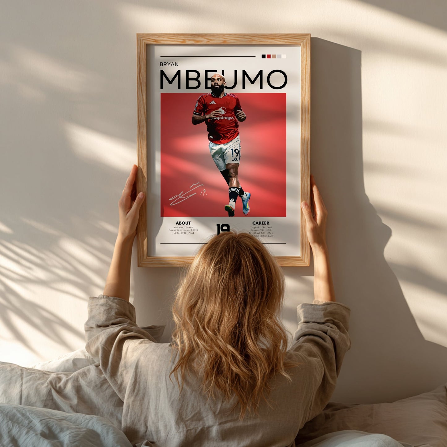 Person hanging a framed poster of a soccer player named Mbeumo on a wall.