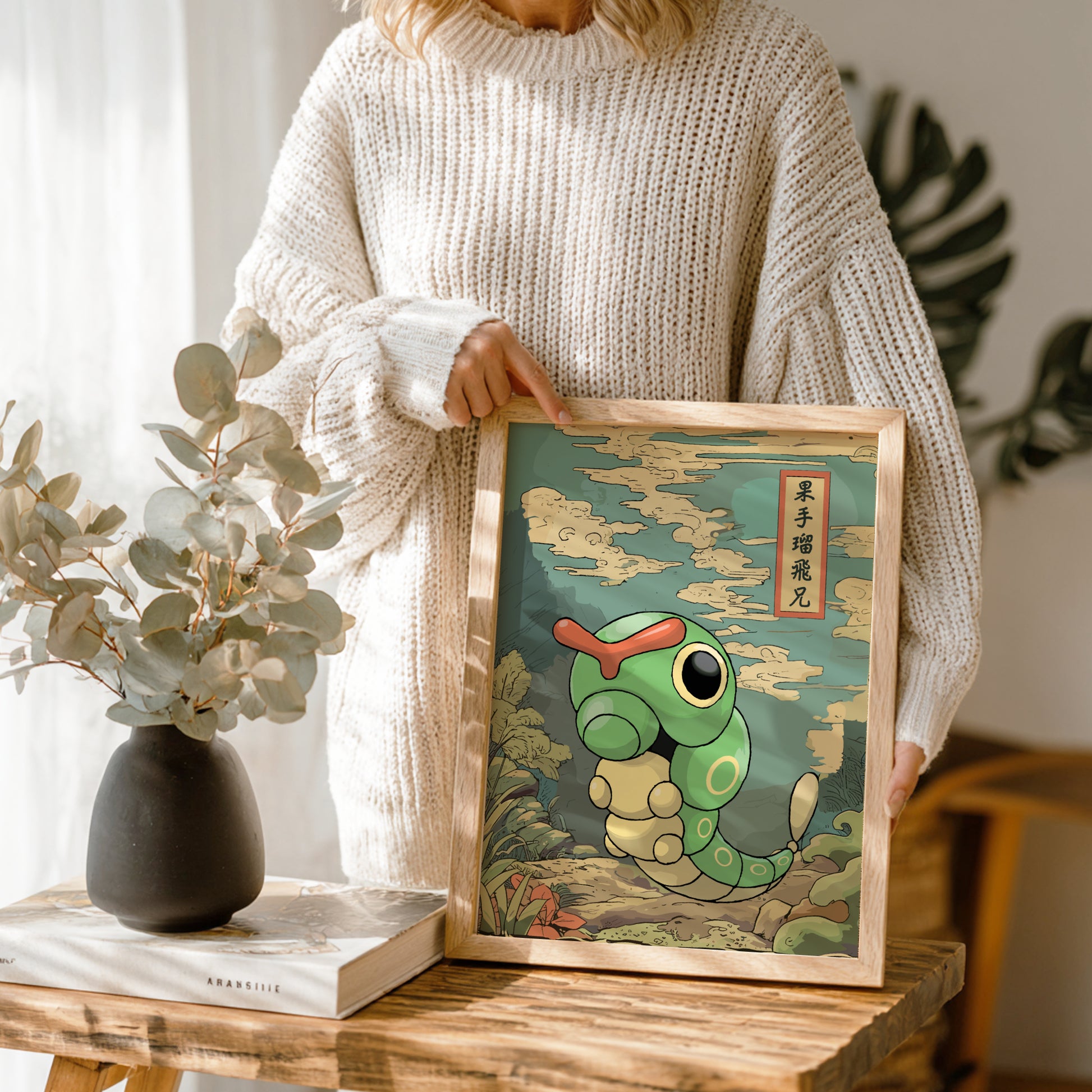 Person holding a framed illustration of Caterpie Pokemon on a wooden table.