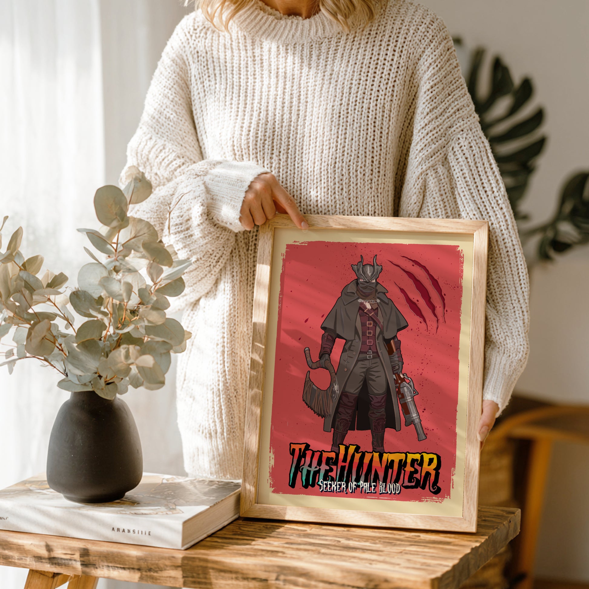Person holding a framed artwork of The Hunter from Bloodborne on a wooden table.