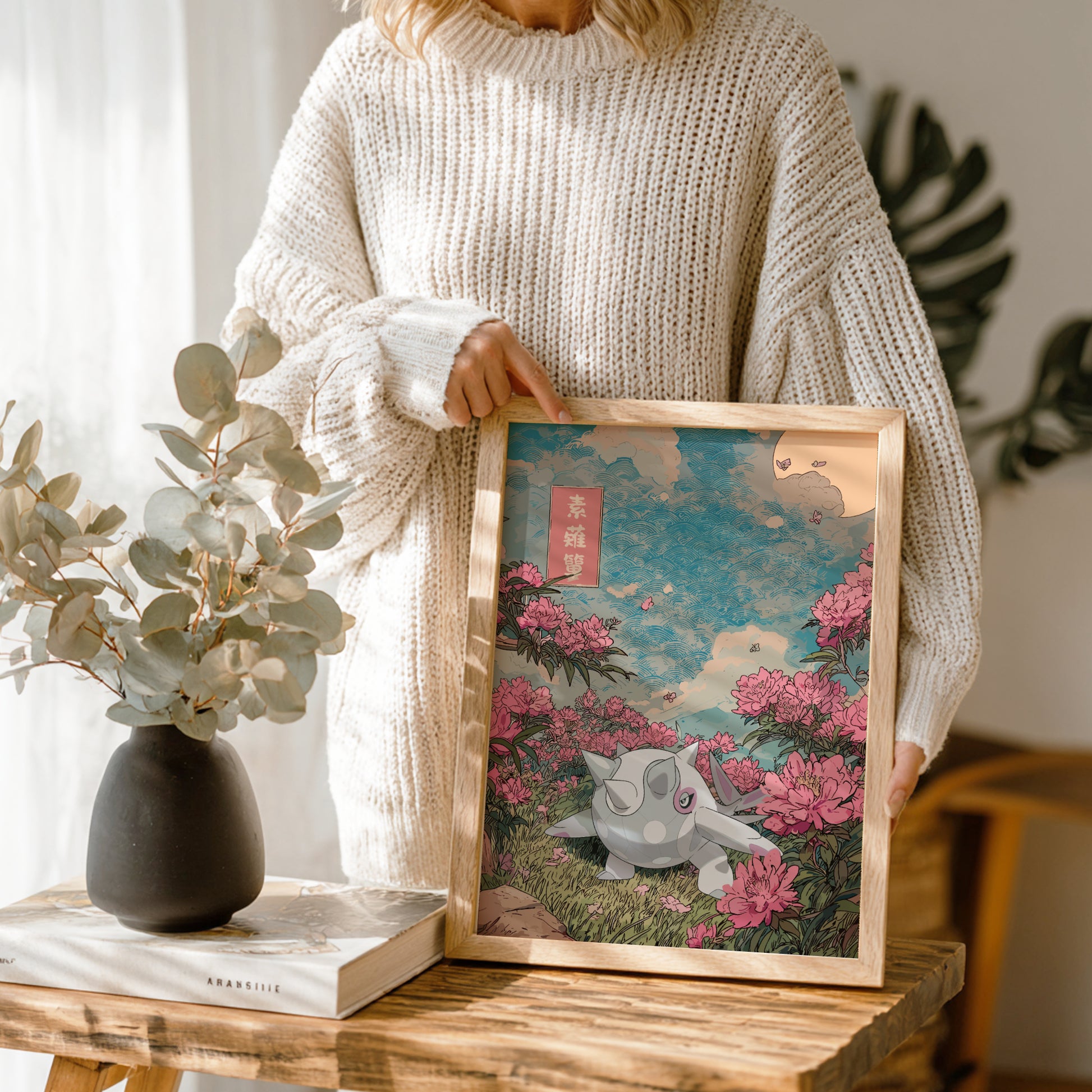 Person holding a framed artwork of Cetitan Pokemon in a floral setting, with a vase and book on a wooden table.