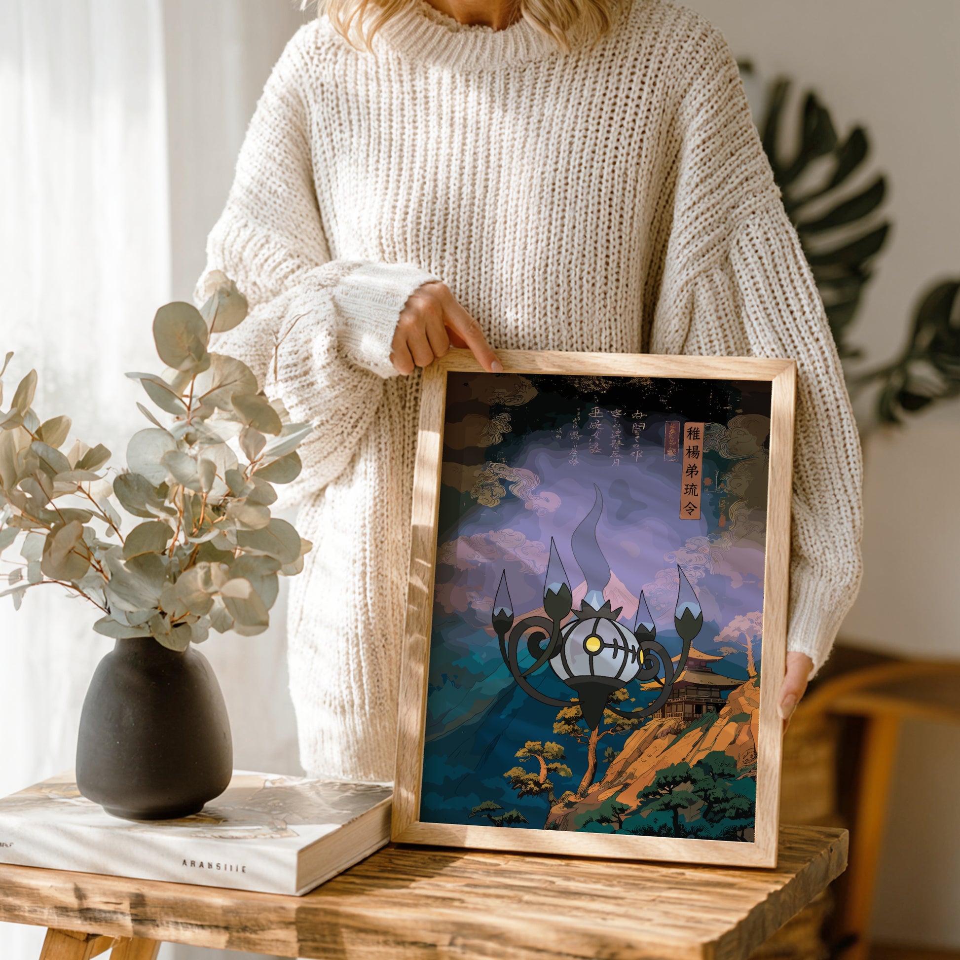 Person holding a framed artwork with Chandelure Pokemon design, standing next to a vase and books on a wooden table.