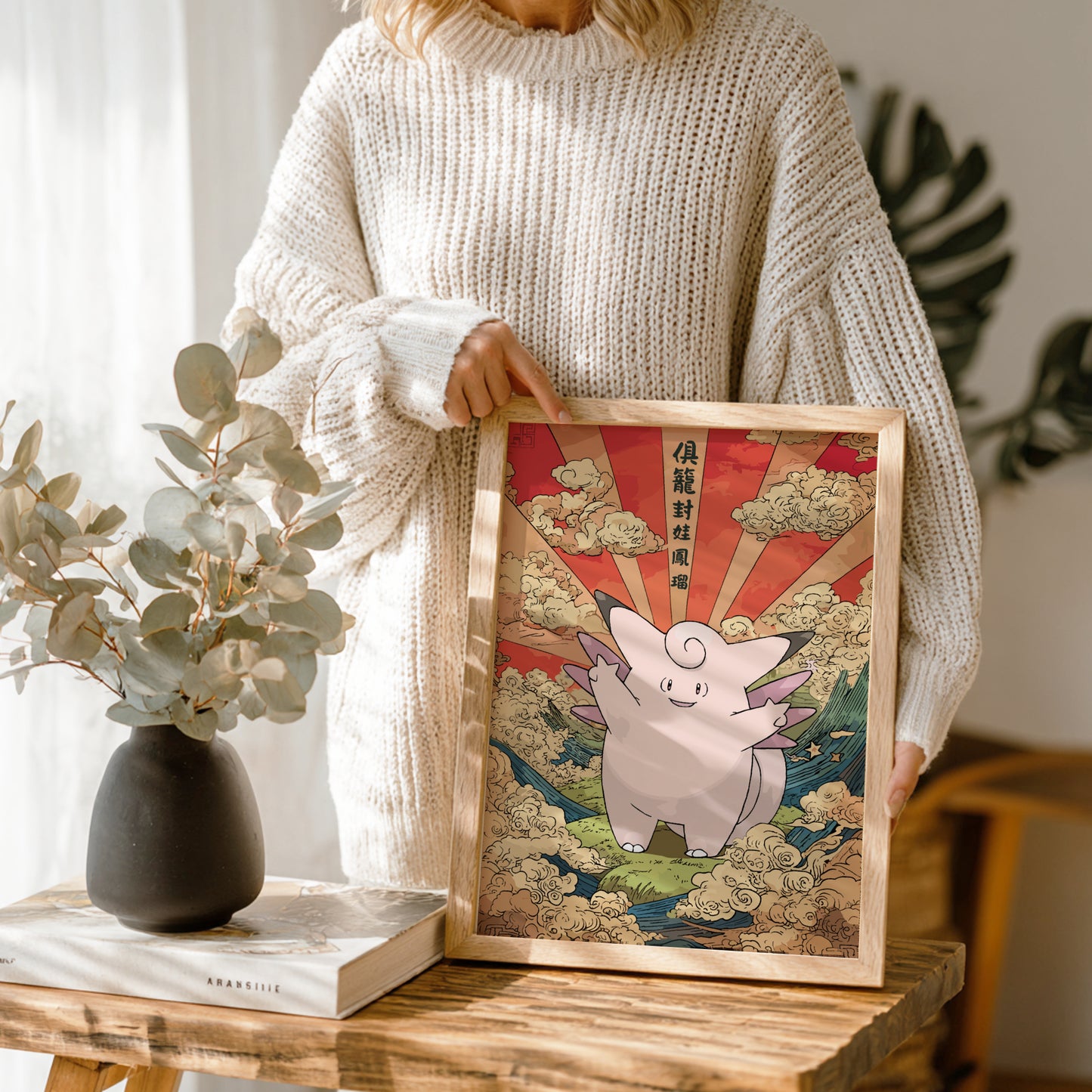 Person holding a framed artwork of Clefable Pokemon with a red sun and clouds in a wooden frame.