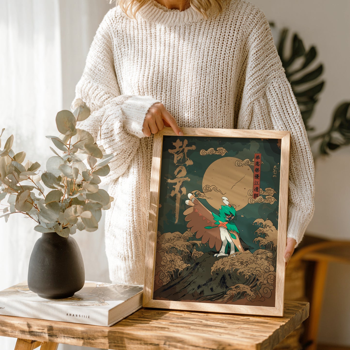 Person holding a framed artwork with a stylized Decidueye Pokemon character and moon design, standing next to a vase and books on a wooden table.