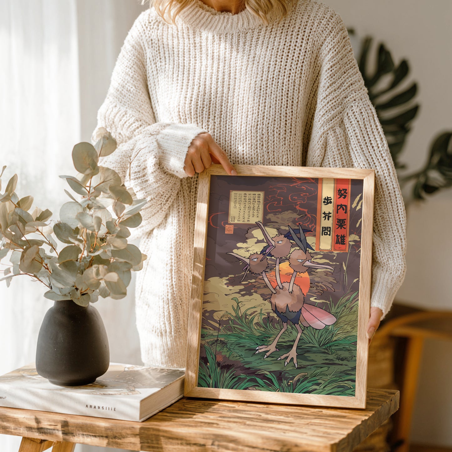 Person holding a framed artwork of Dodrio Pokemon with traditional Japanese text, standing in a room with plants and a vase.
