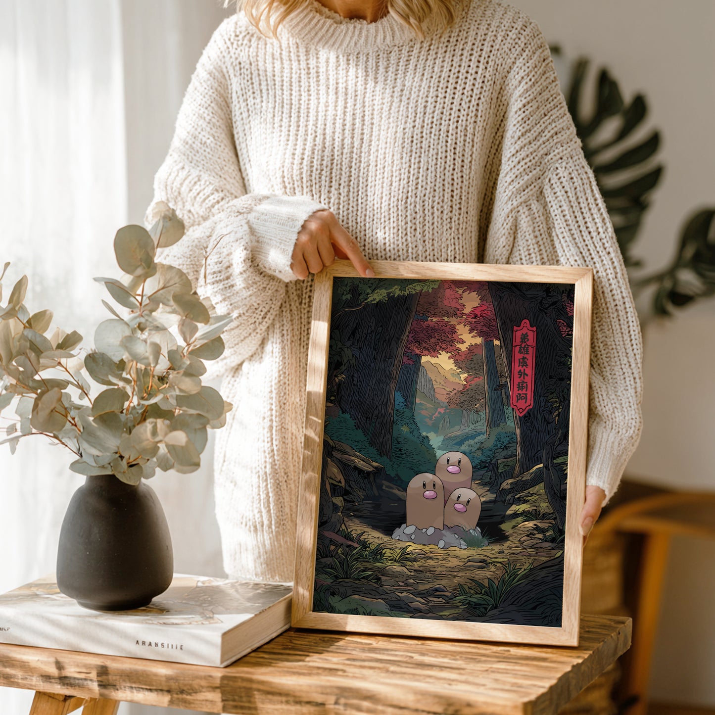 Person holding a framed artwork of Dugtrio Pokemon character in a forest scene, with a vase and book on a table.
