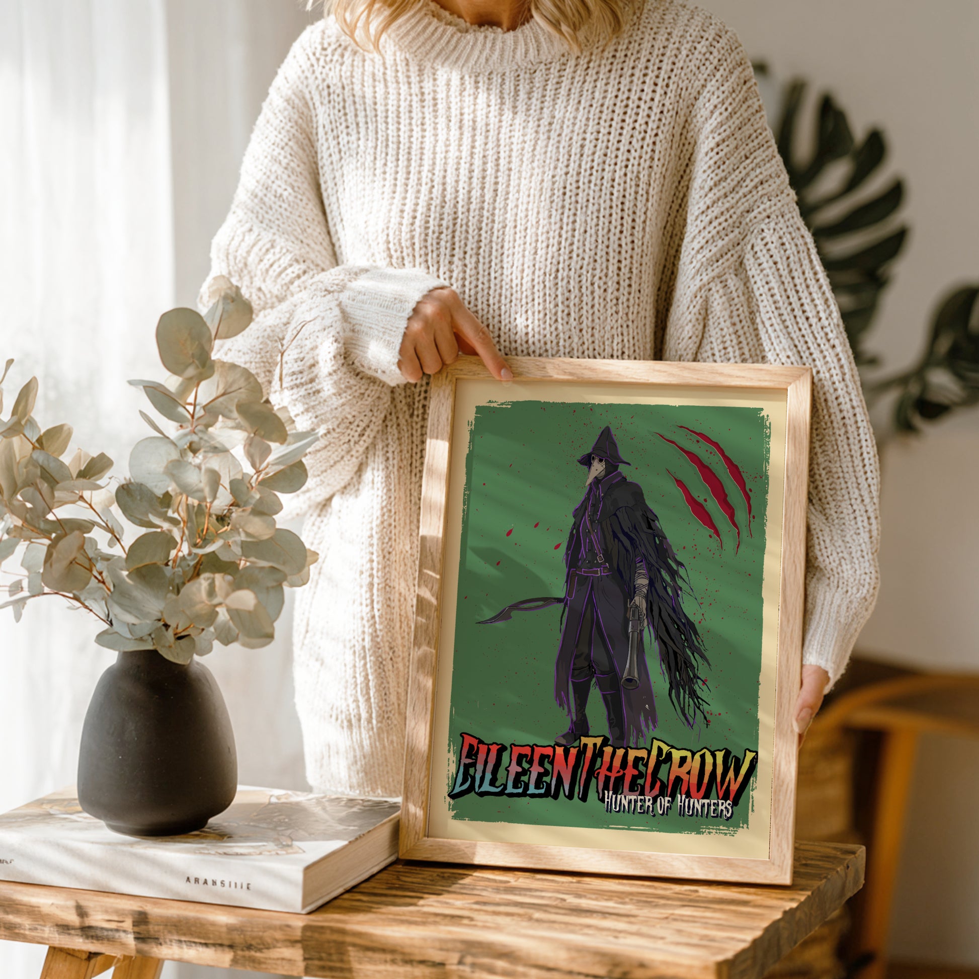 Person holding a framed artwork of Eileen The Crow from Bloodborne on a wooden table.
