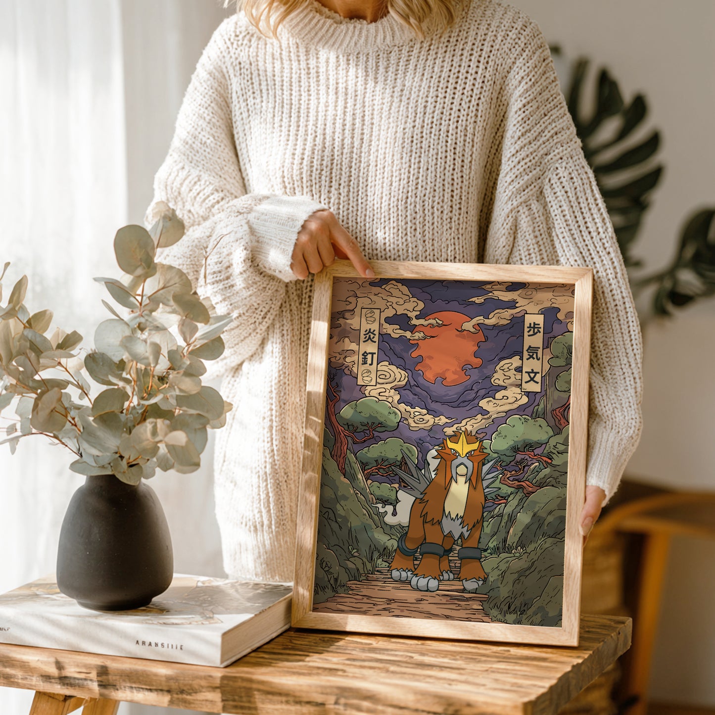 Person holding a framed artwork of Entei Pokemon with a mountain background, standing in a room with plants and a vase.