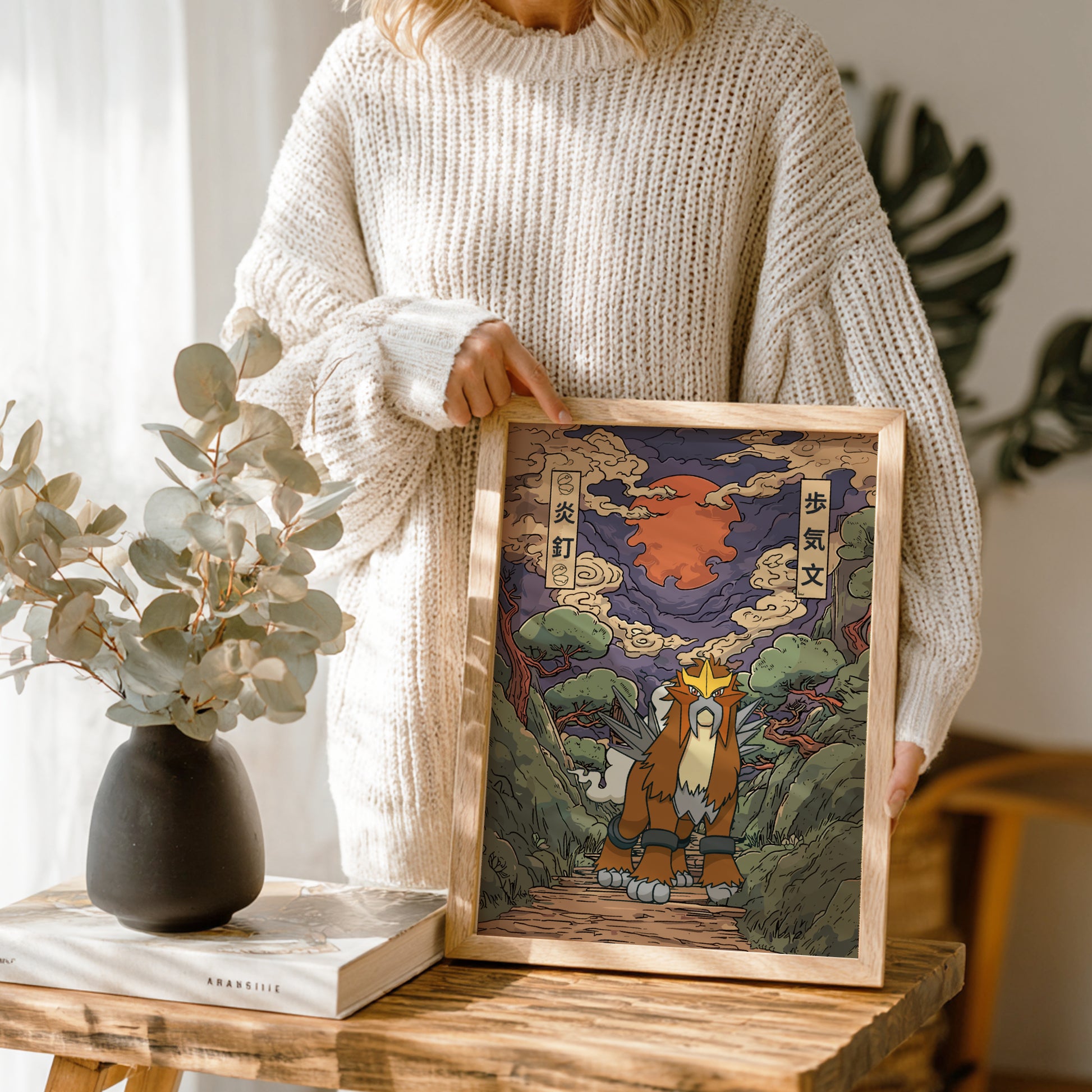 Person holding a framed artwork of Entei Pokemon with a mountain background, standing in a room with plants and a vase.