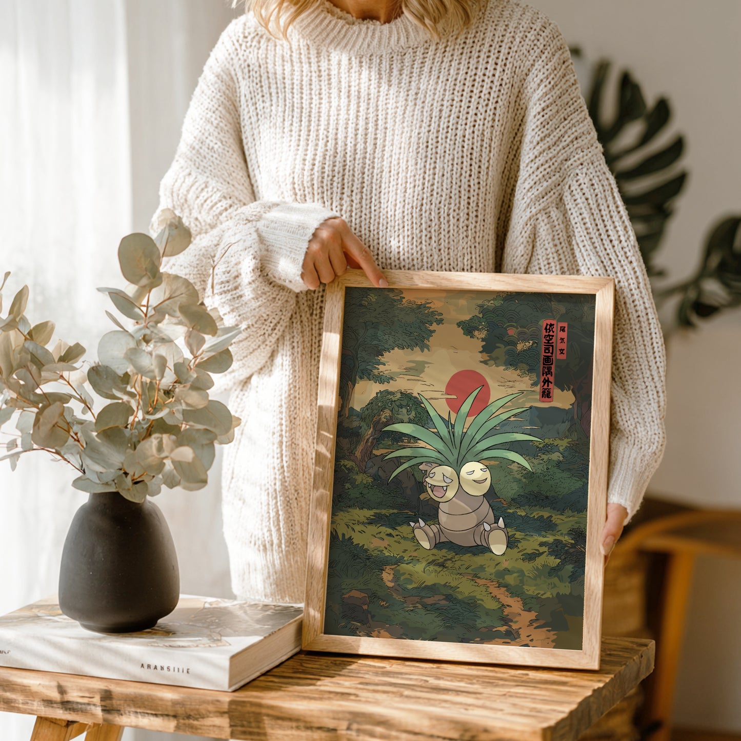 Person holding a framed artwork of Exeggutor Pokemon on a wooden table with a vase and book in the background.
