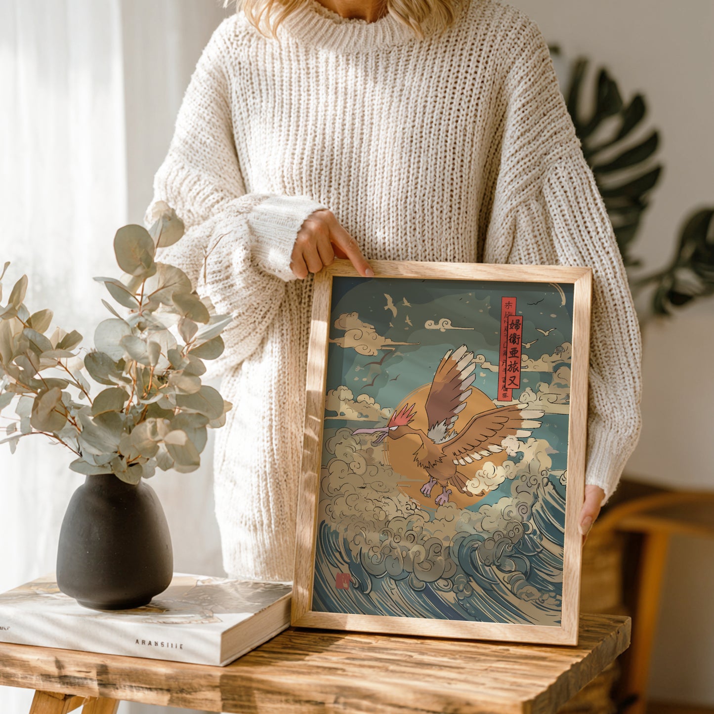 Person holding a framed artwork of Fearow Pokemon on a wave, with a vase and book on a wooden table.