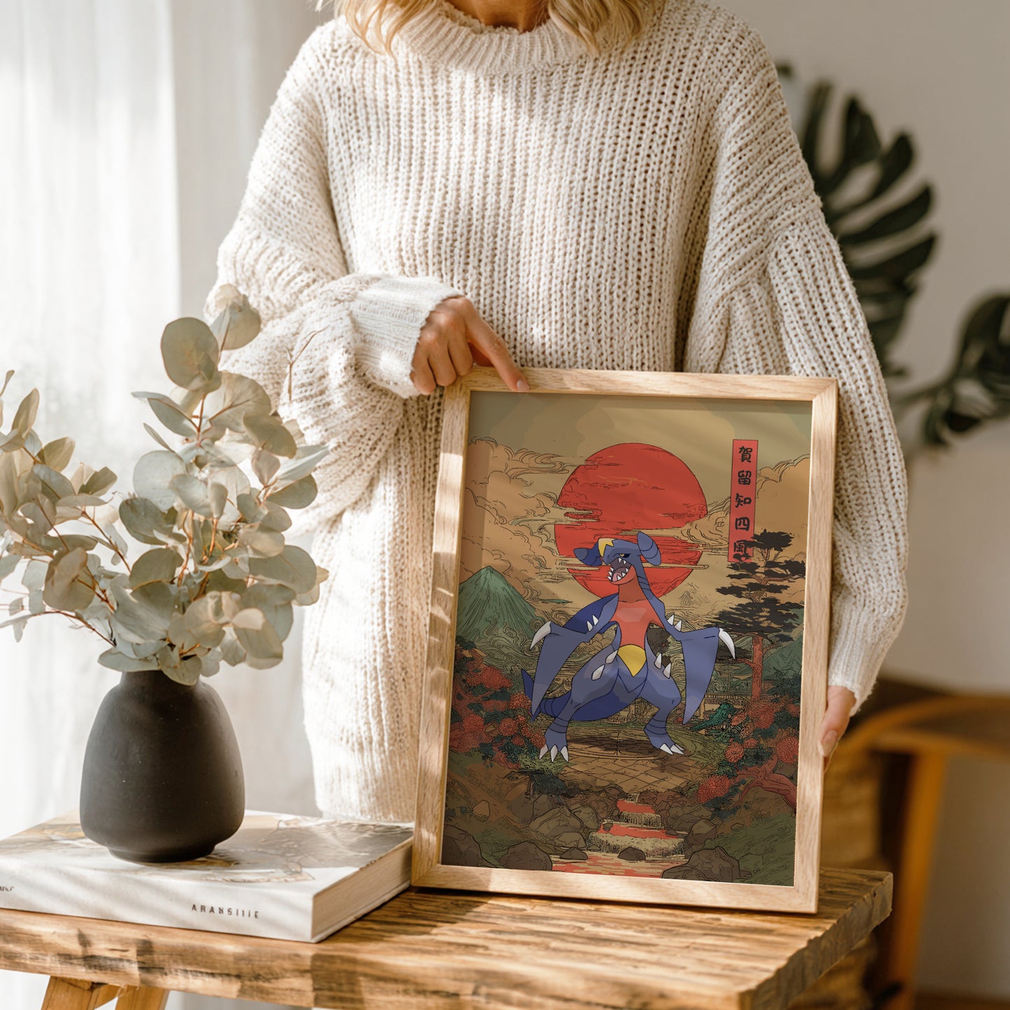 Person holding a framed artwork with a stylized depiction of Garchomp Pokemon  and a red sun, standing in a room with a vase and plant.