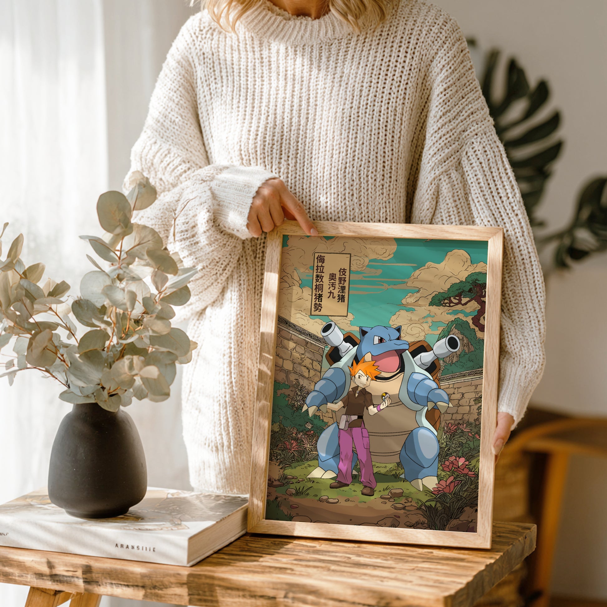 Person holding a framed artwork of Gary Oak (Blue) and Blastoise characters on a wooden table with a vase and book in the background.