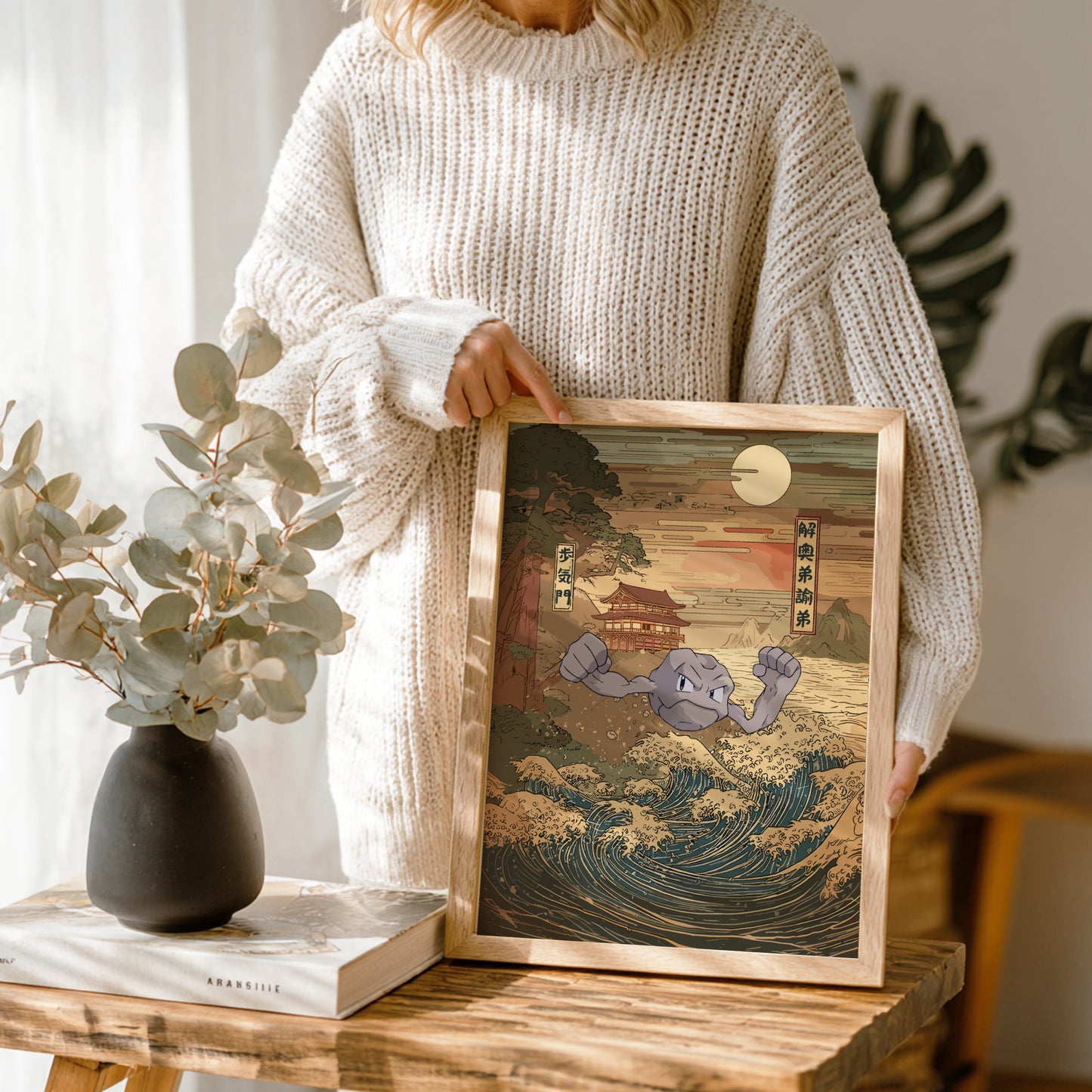 Person holding a framed artwork of Geodude Pokemon on a wave with a vase and book in the background