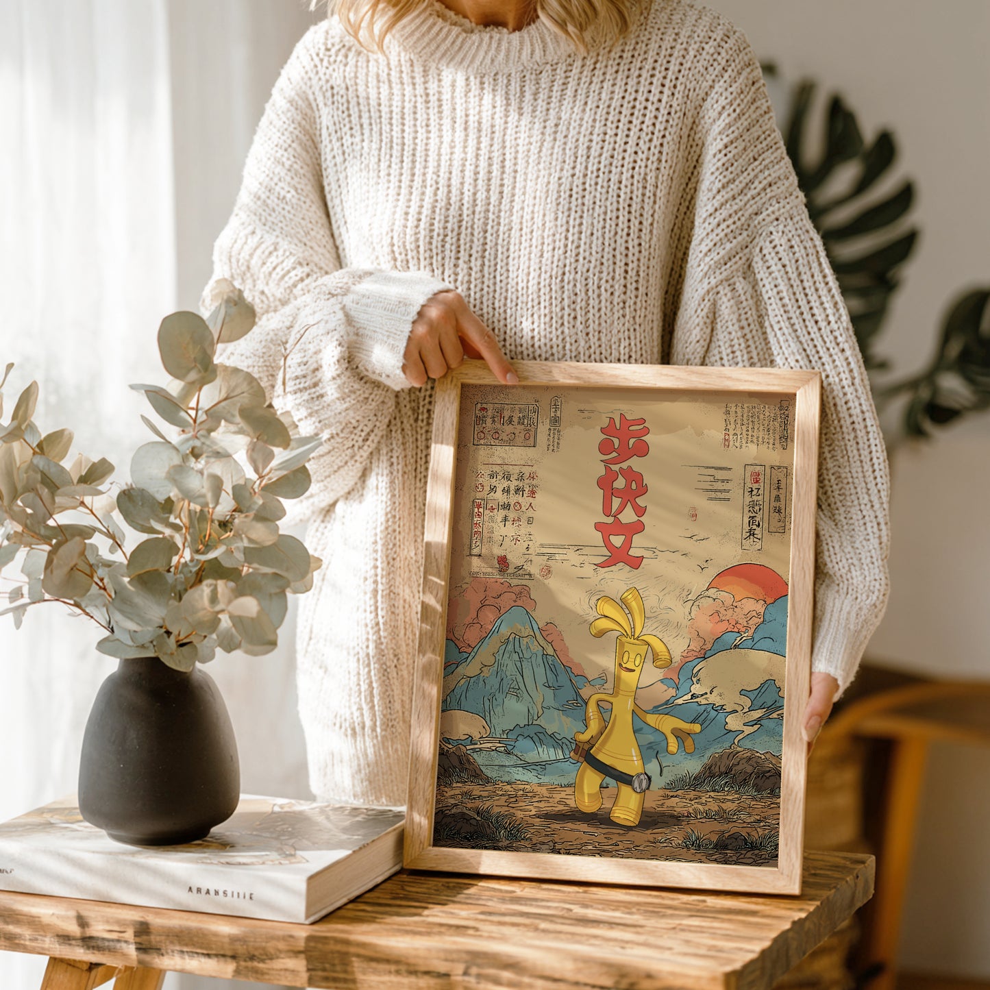 Person holding a framed artwork with a Gholdengo Pokemon character and mountain landscape, standing in a room with plants and a vase.