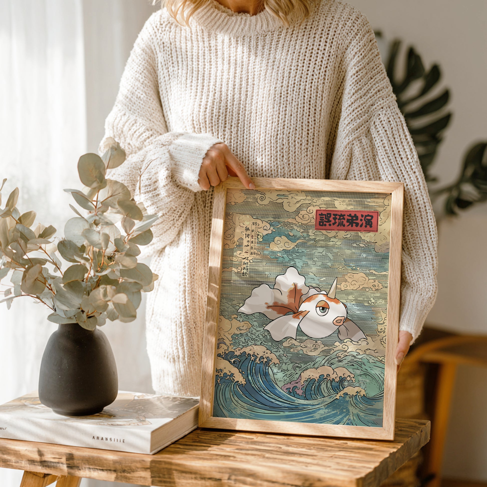 Person holding a framed artwork of Goldeen Pokemon in waves on a wooden table with a vase and book in the background.