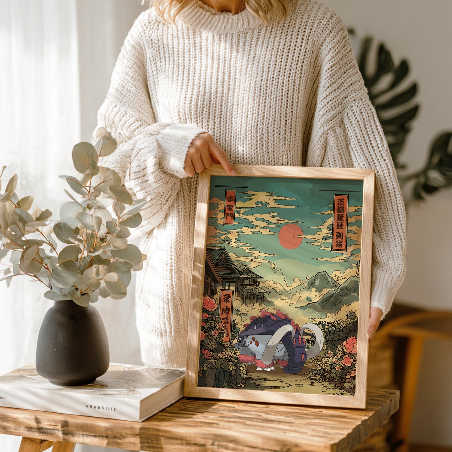 Person holding a framed artwork with Great Tusk Pokemon and a scenic design, standing next to a vase and books on a wooden table.
