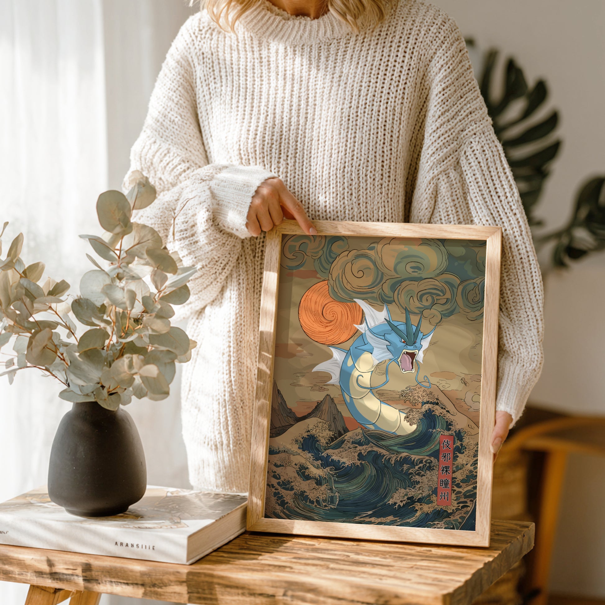 Person holding a framed artwork of Gyarados Pokemon in a wooden frame, with a vase and book on a table in the background.