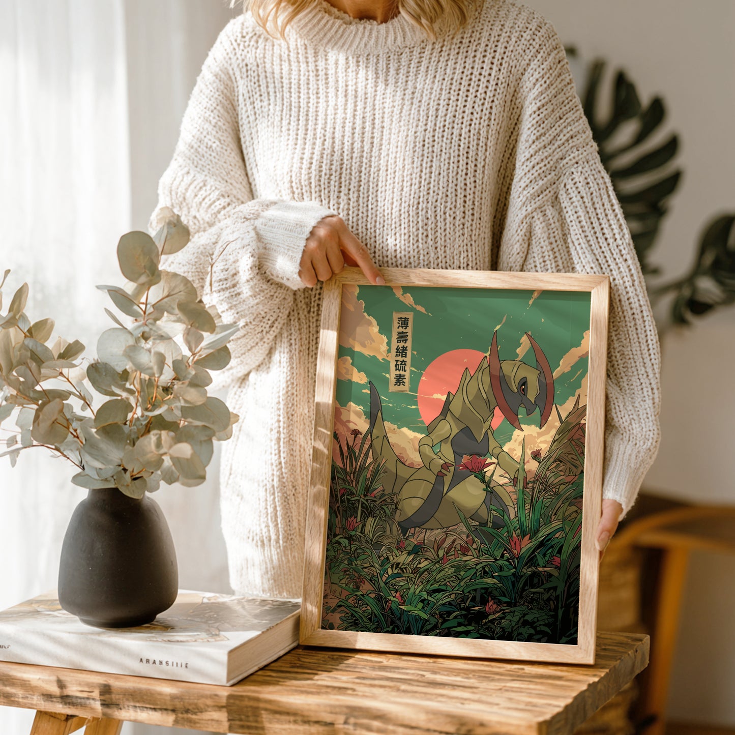 Person holding a framed artwork of Haxorus Pokemon in a forest with a vase and book on a table.