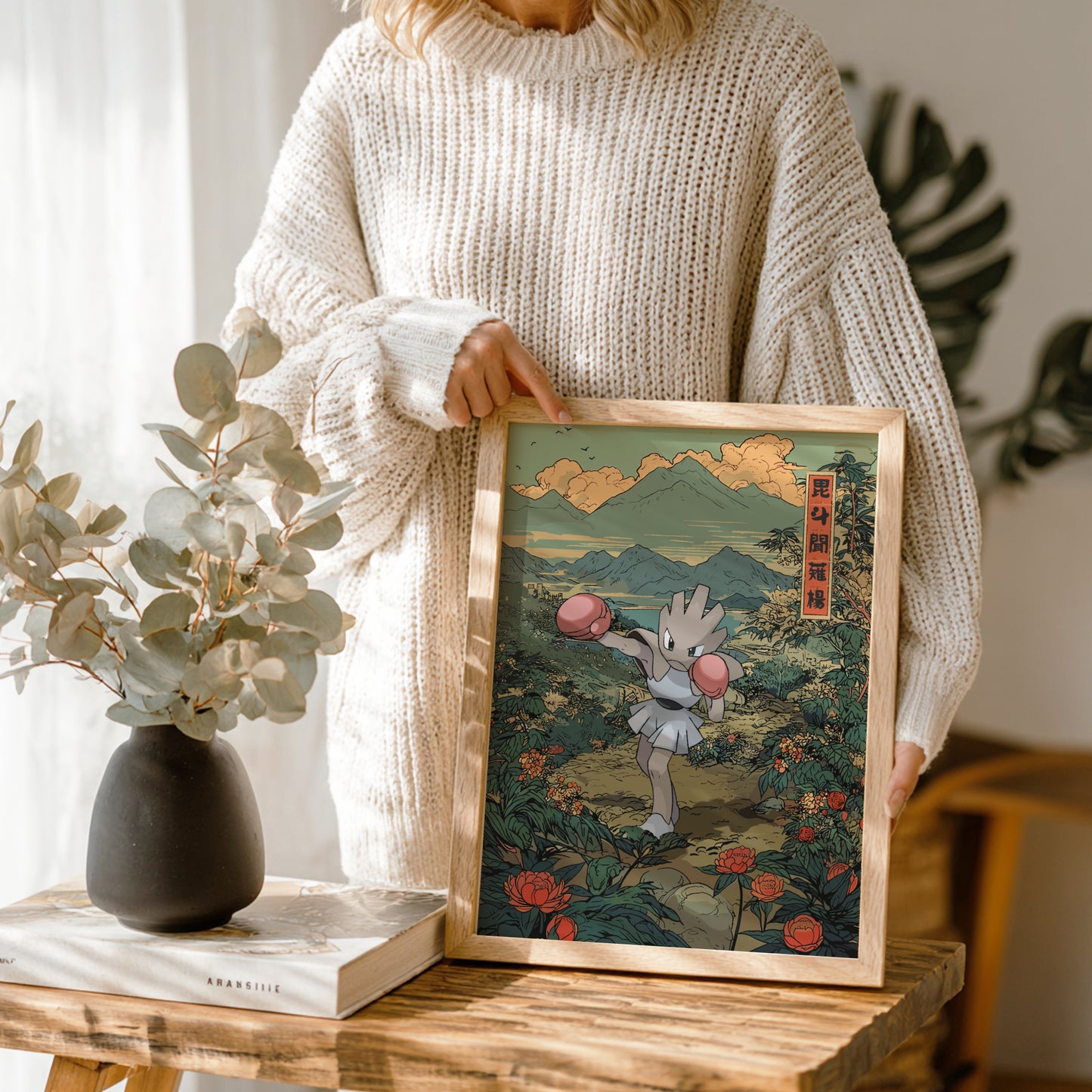 Person holding a framed artwork of Hitmonchan Pokemon in a scenic setting, with a vase and books on a table.
