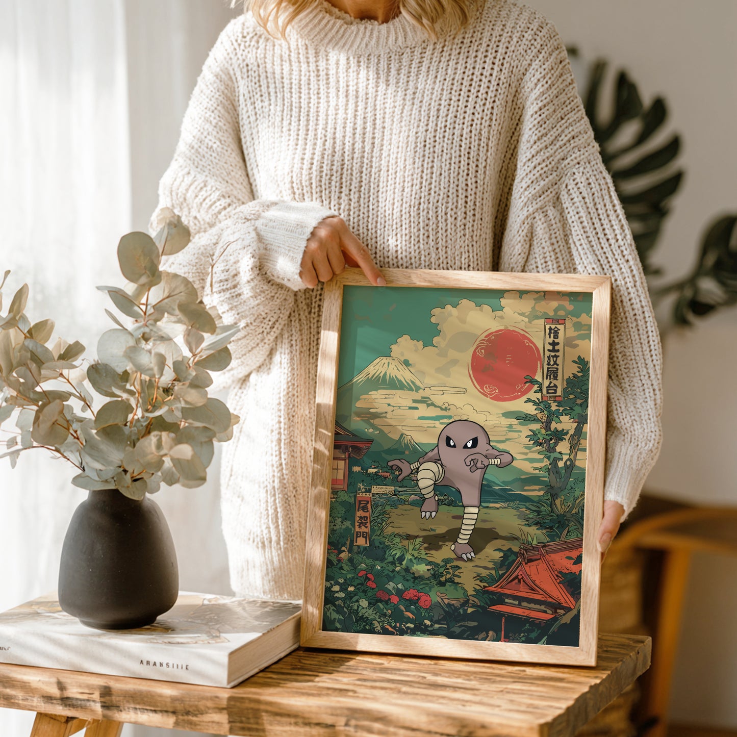 Person holding a framed artwork with a mountain and sun and Hitmonlee Pokemon design, standing next to a vase and books on a wooden table.