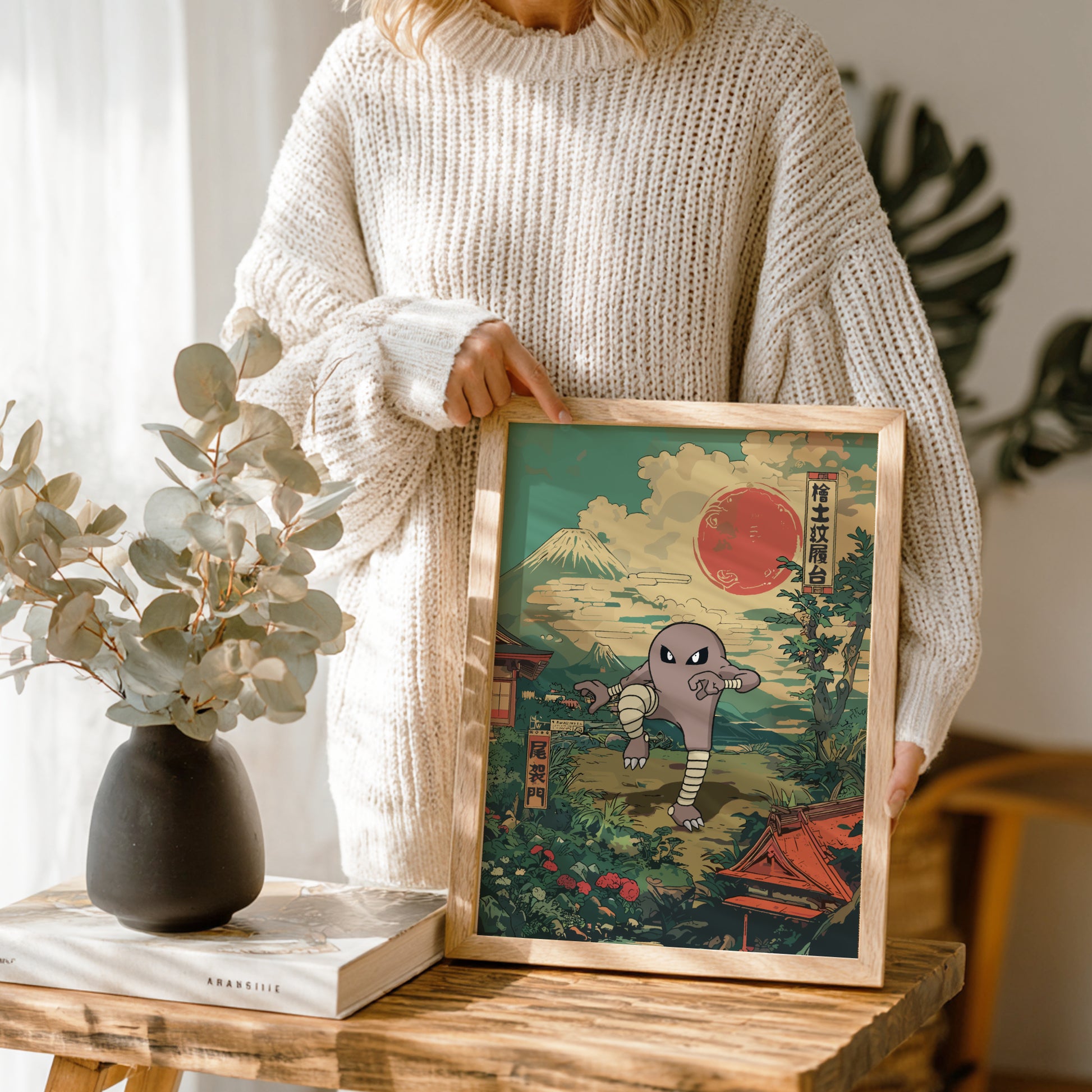 Person holding a framed artwork with a mountain and sun and Hitmonlee Pokemon design, standing next to a vase and books on a wooden table.