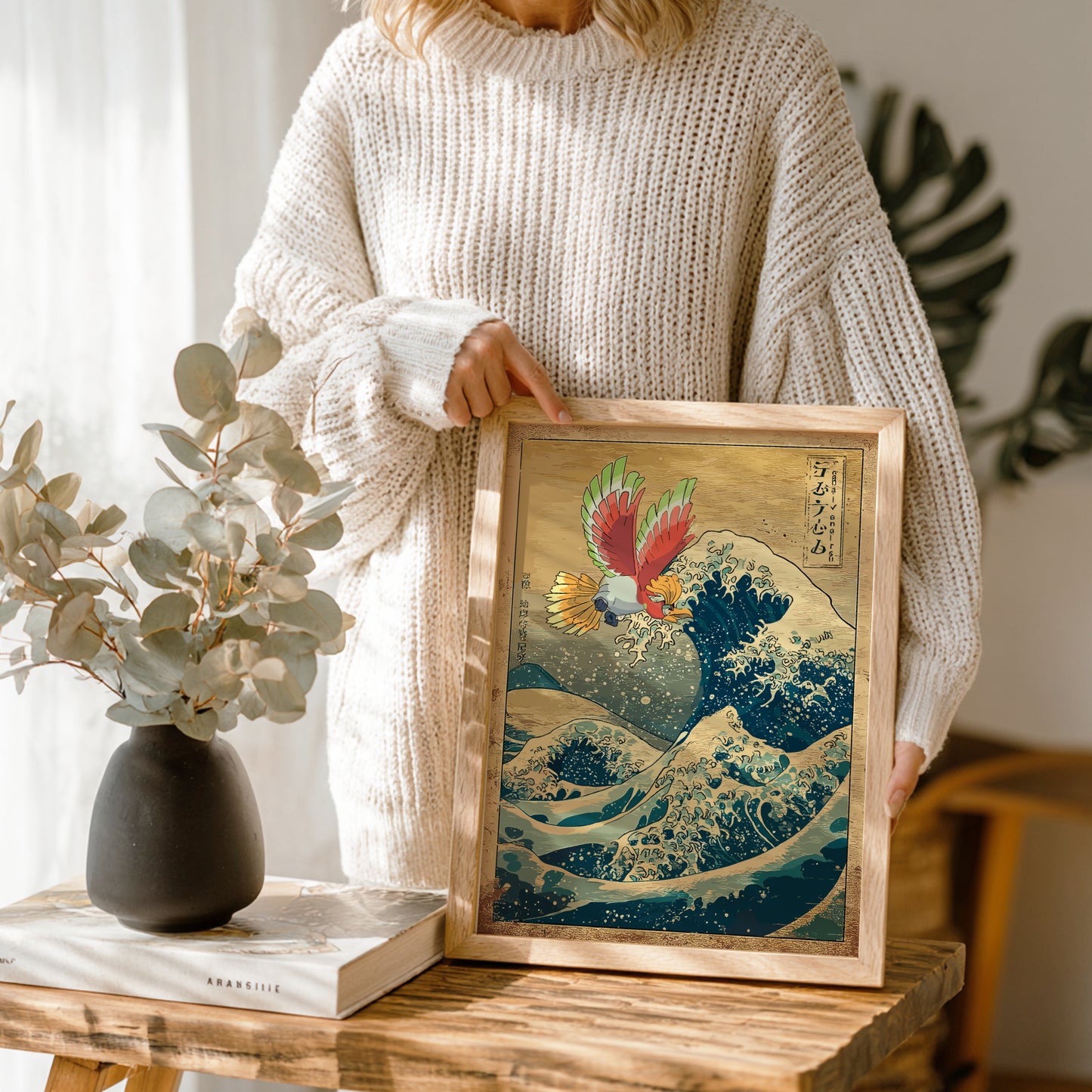 Person holding a framed artwork of Ho-Oh Pokemon on a wave, standing next to a vase with greenery.