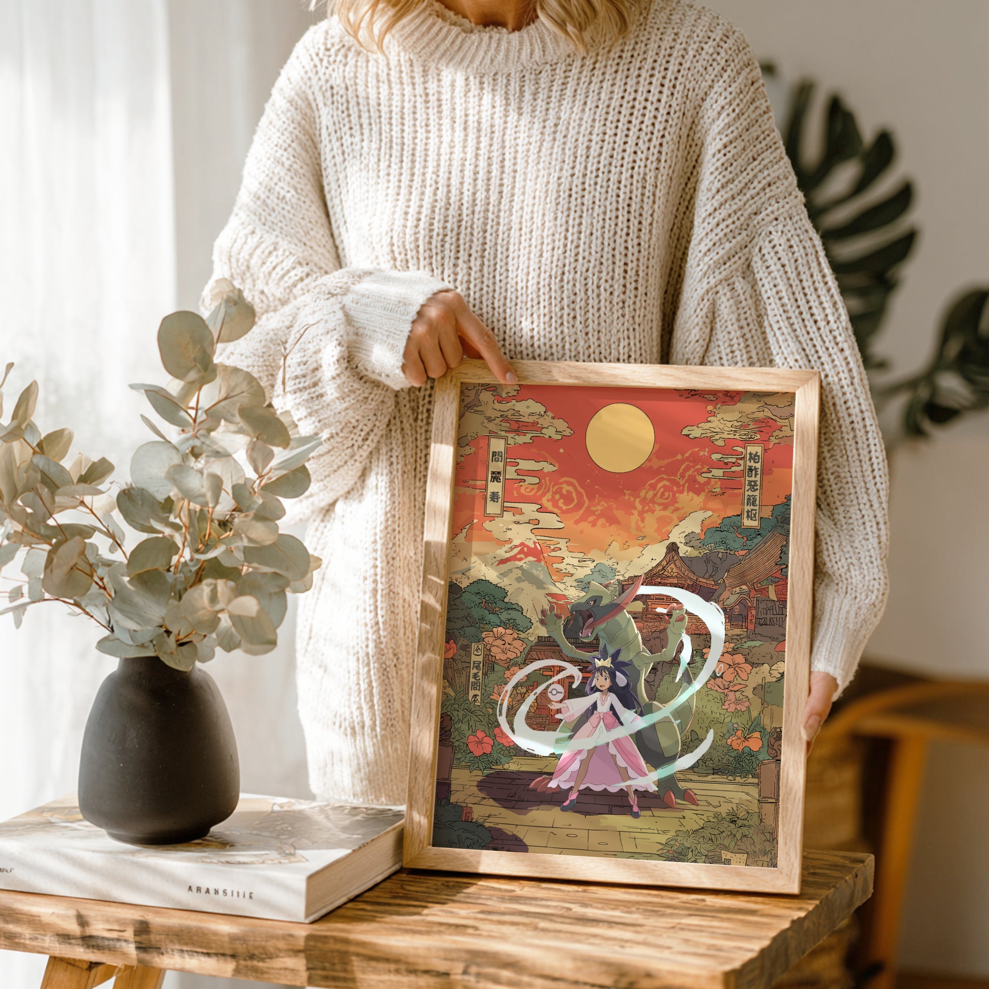 Person holding a framed artwork with Iris and Haxorus Pokemon design, standing next to a vase and books on a wooden table.