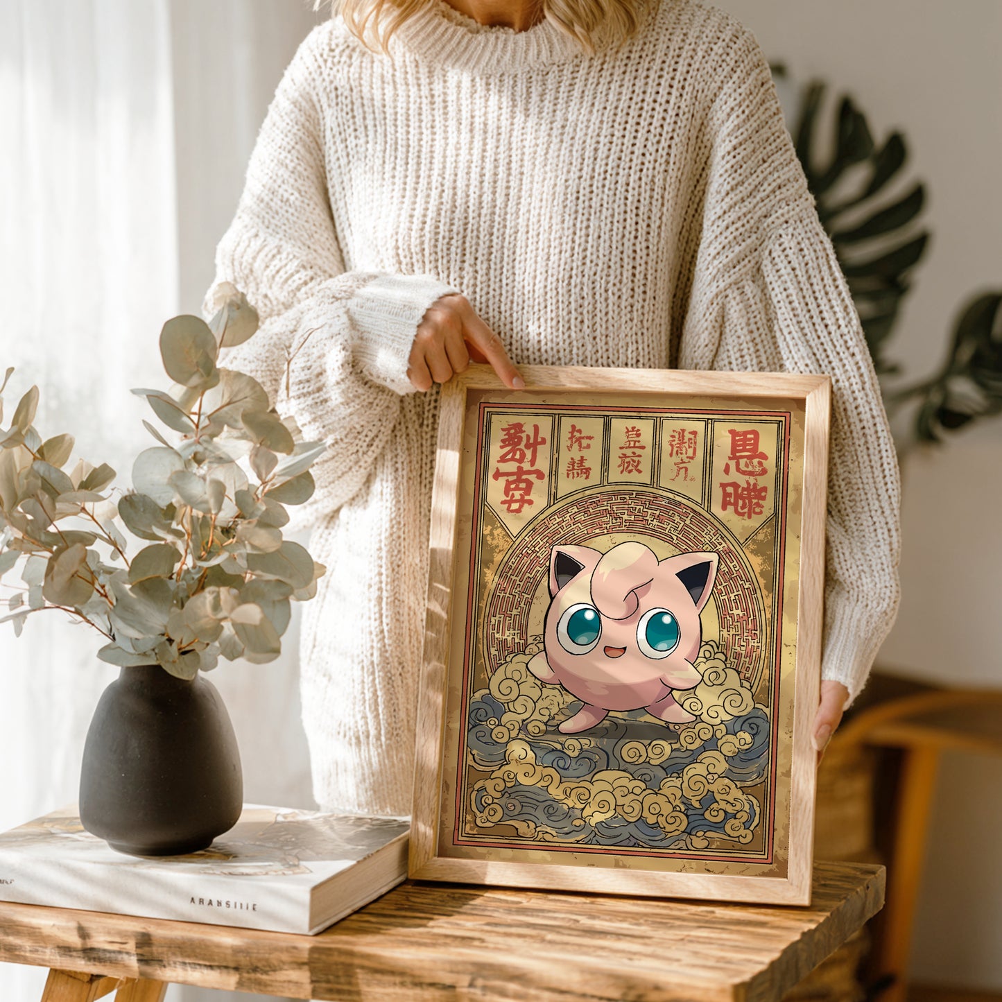 Person holding a framed illustration of Jigglypuff Pokemon character with decorative elements on a wooden table.