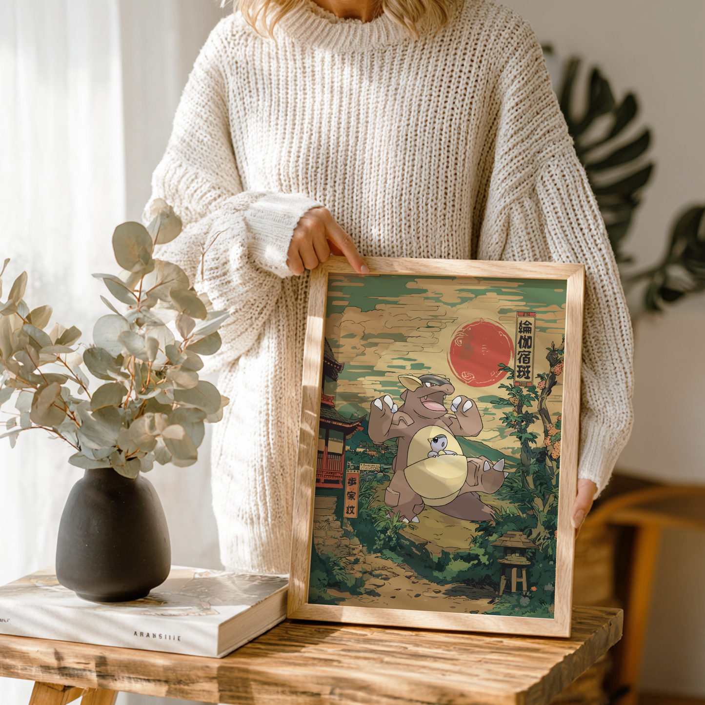 Person holding a framed artwork of Kangaskhan Pokemon in a traditional Japanese setting on a wooden table.