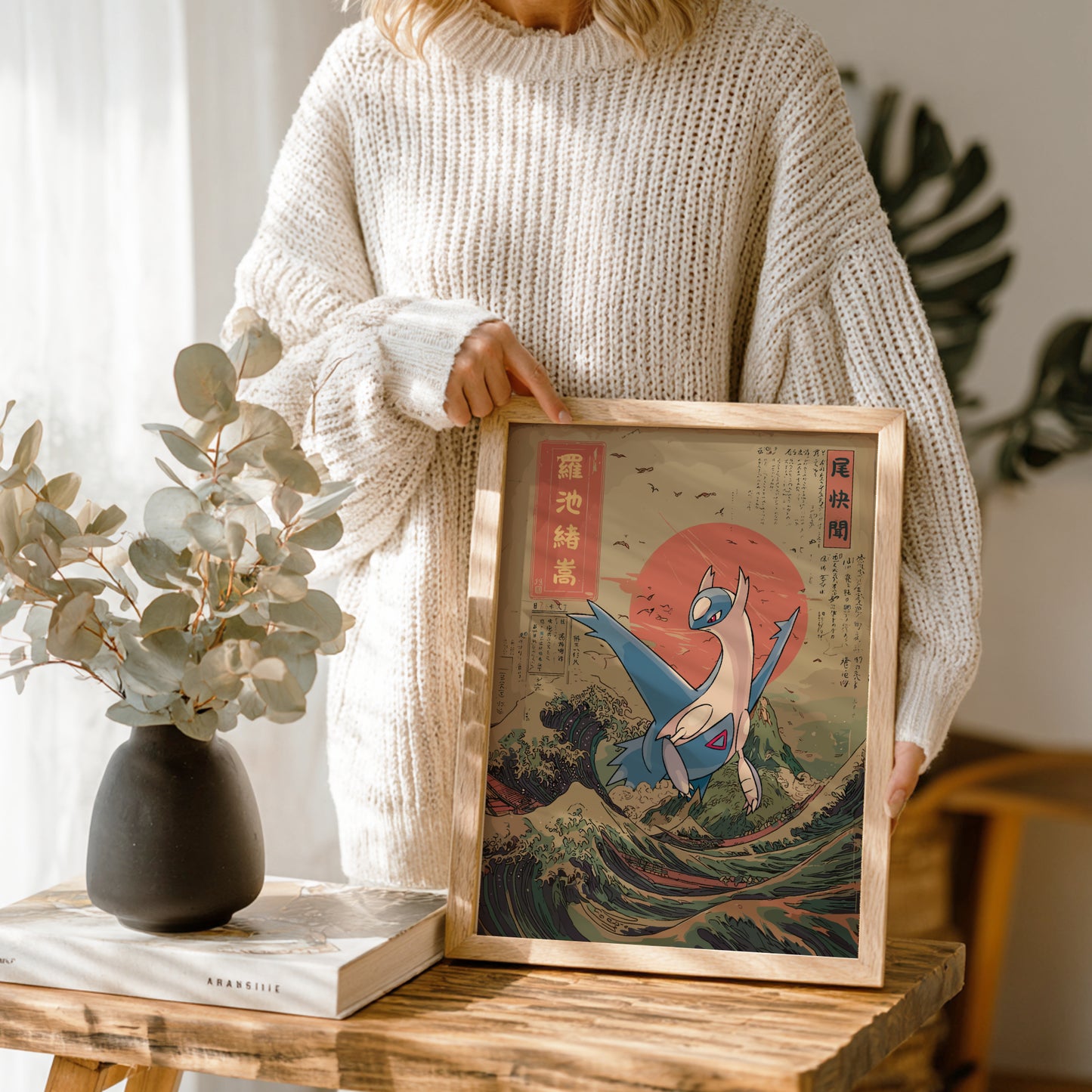 Person holding a framed artwork of Latios Pokemon on a wave with a vase and book in the background