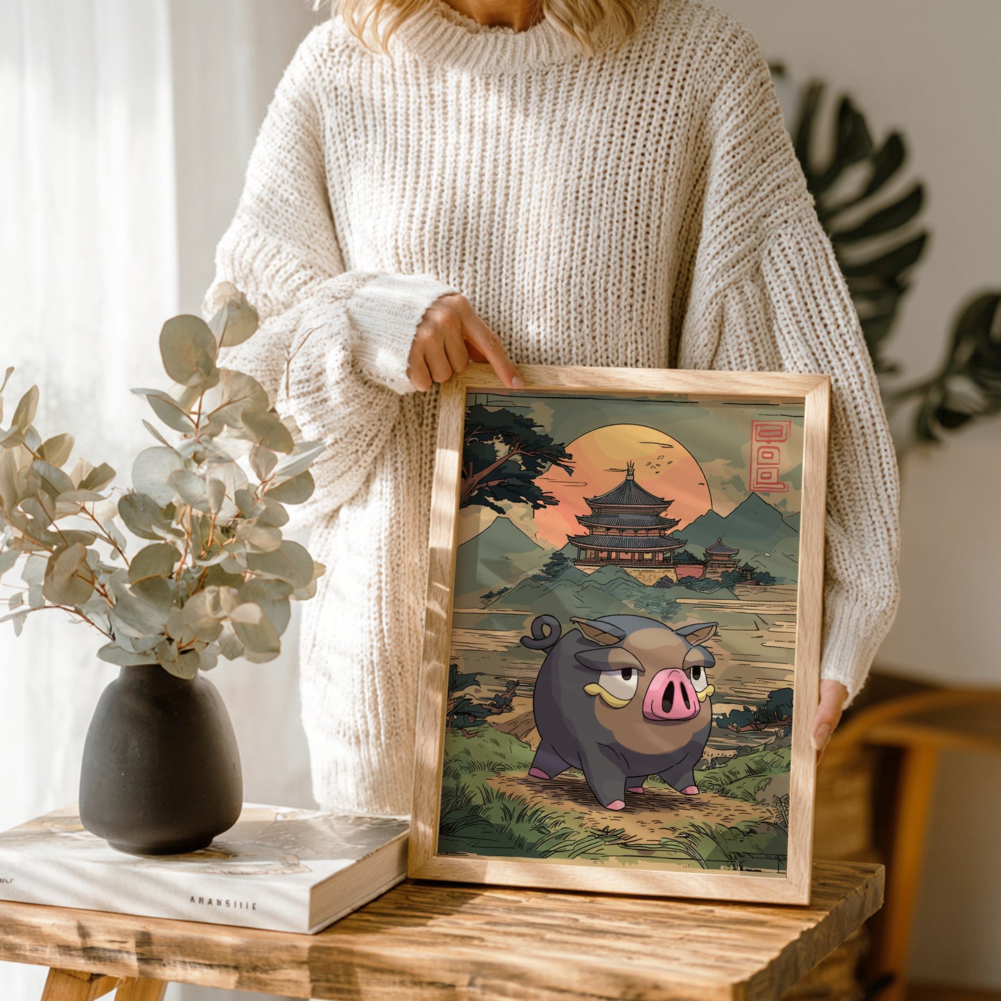 Person holding a framed illustration of Lechonk Pokemon in a scenic setting, with a vase and books on a table.