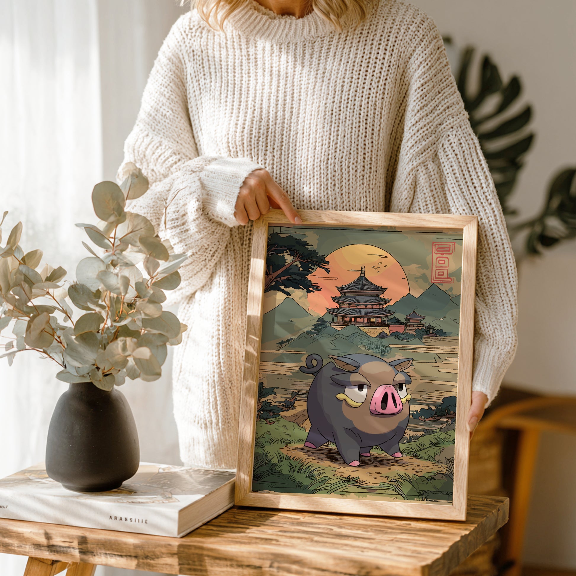 Person holding a framed illustration of Lechonk Pokemon in a scenic setting, with a vase and books on a table.