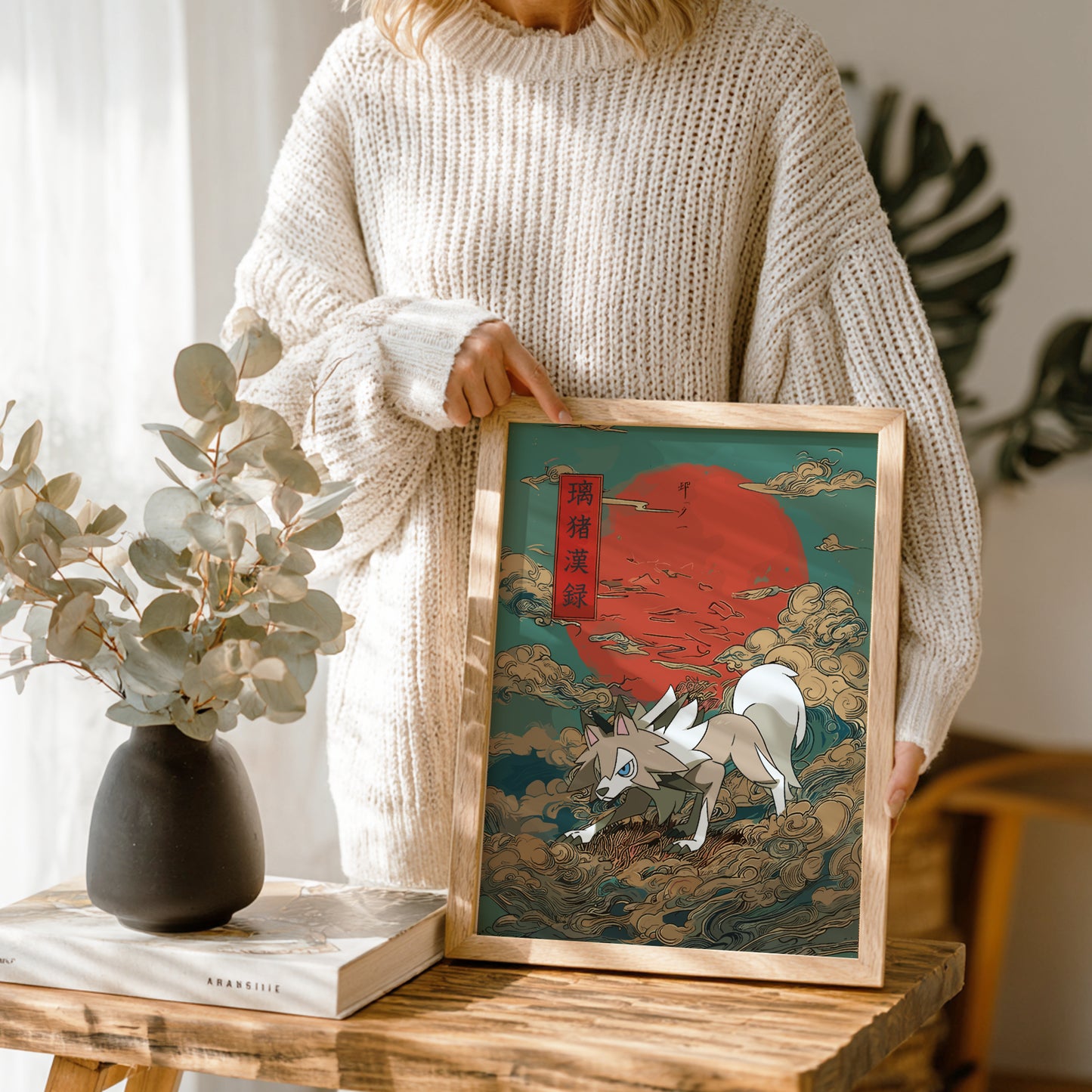 Person holding a framed artwork of Lycanroc Pokemon and a red sun in a room with a vase and book on a table.