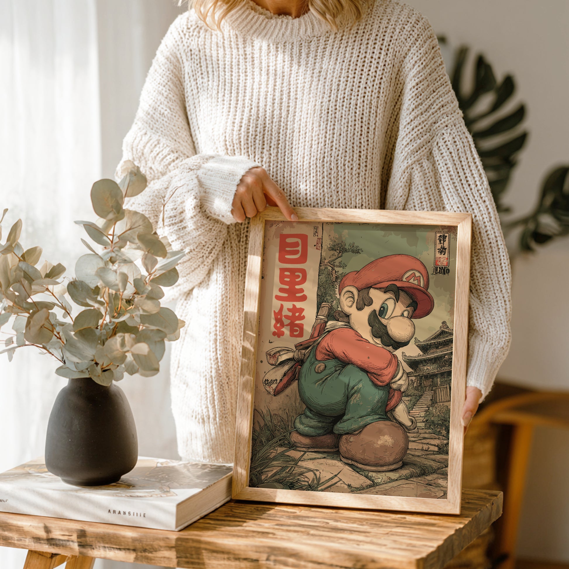 Person holding a framed illustration of Super Mario, with a vase and books on a wooden table.