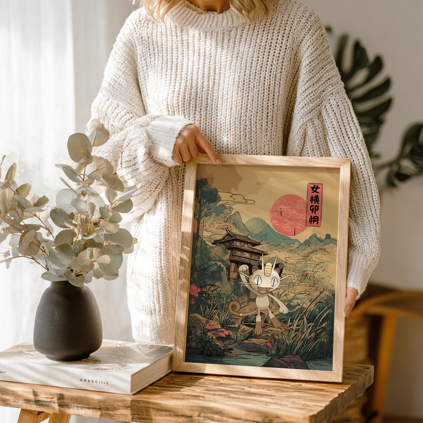 Person holding a framed artwork of a traditional scene with a mountain and a red sun of Meowth Pokemon.