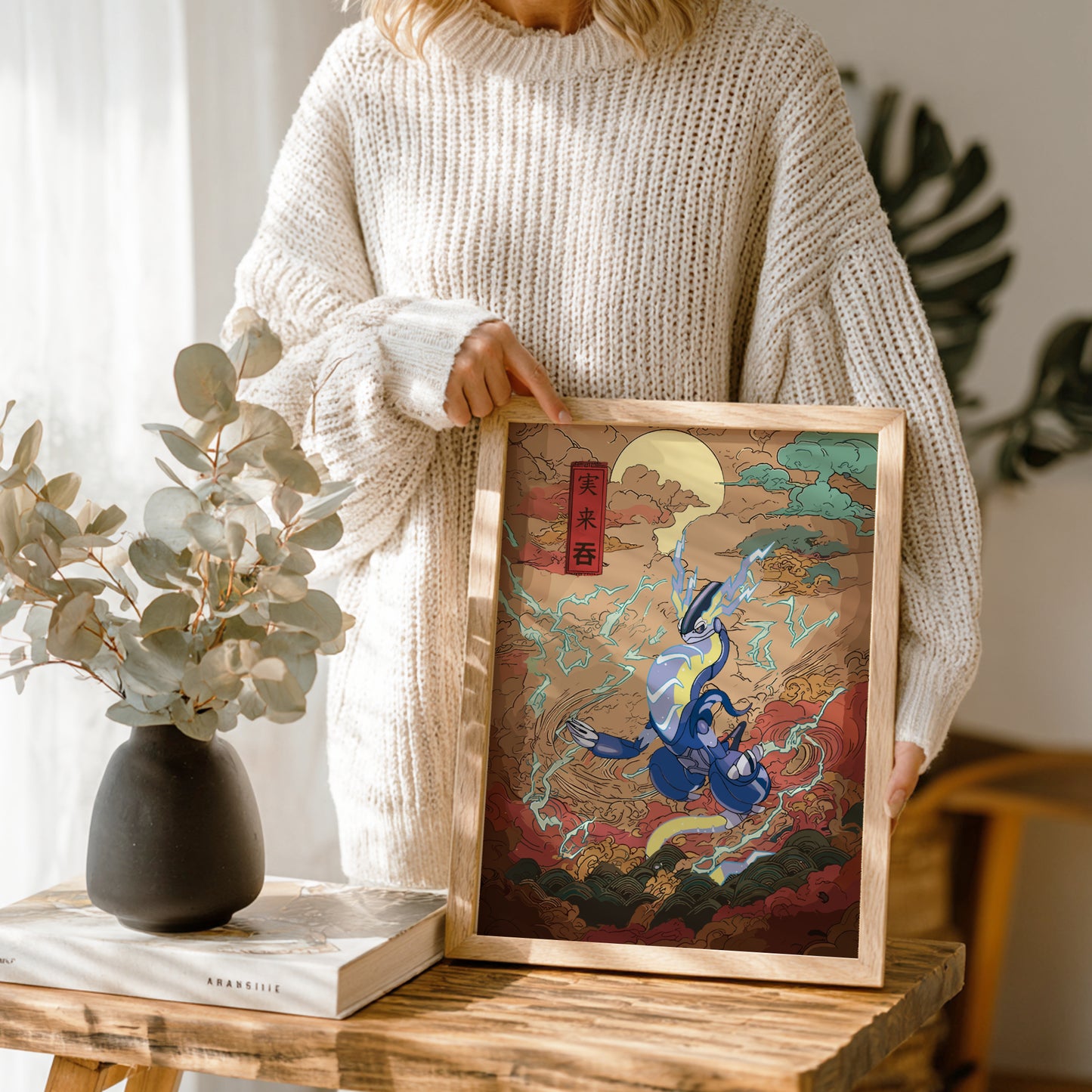 Person holding a framed artwork with a colorful design of Miraidon Pokemon, standing next to a vase and books on a wooden table.