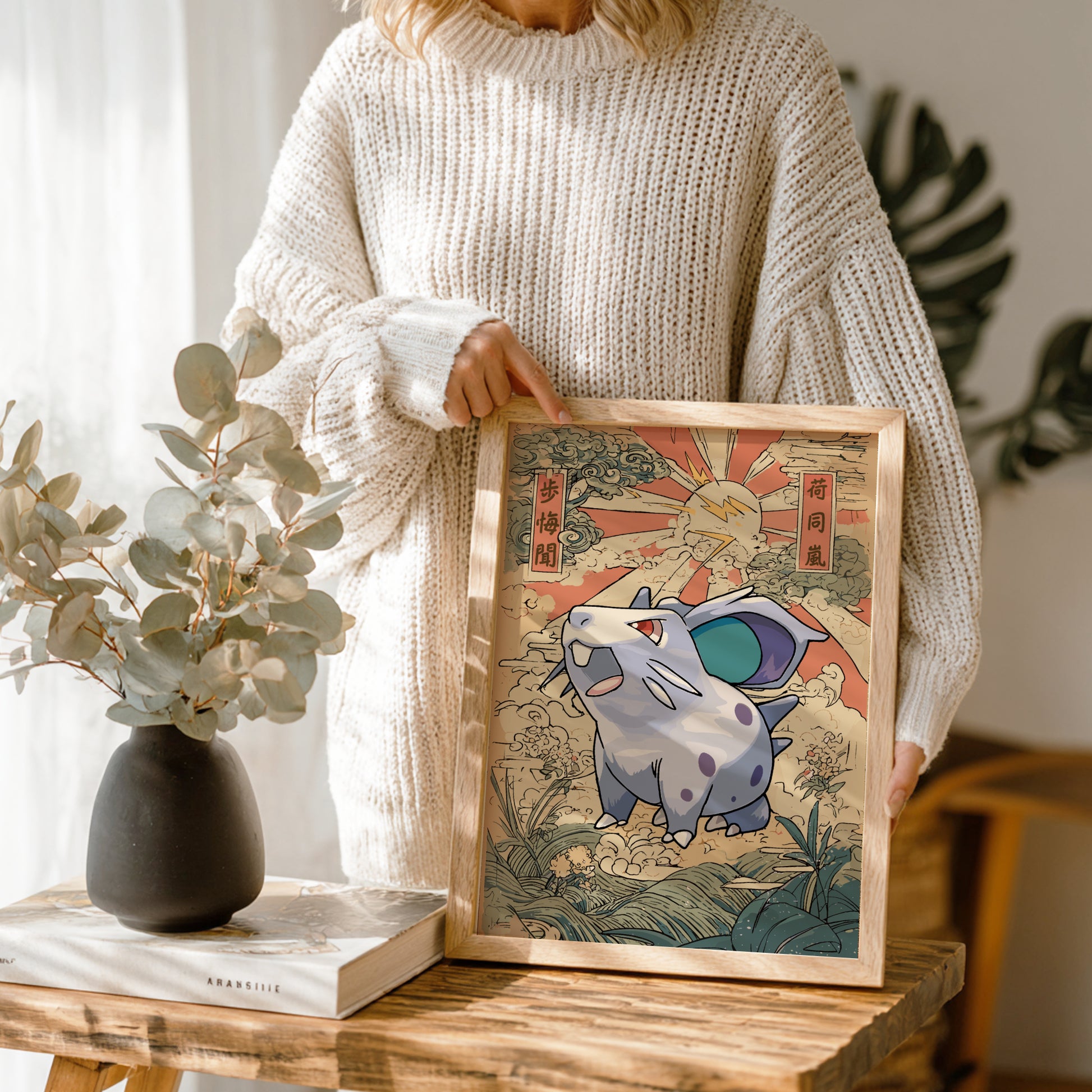Person holding a framed illustration of Nidoran Pokemon character on a wooden table with a vase and book in the background.