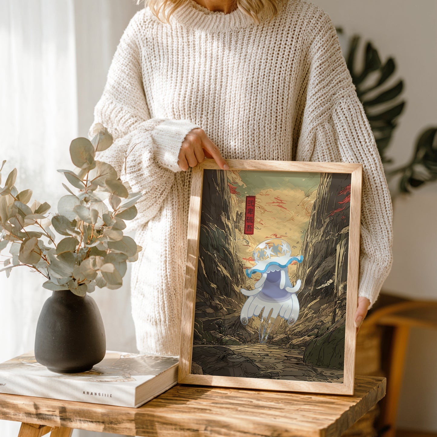 Person holding a framed artwork of Nihilego Pokemon on a wooden table with a vase and book in the background