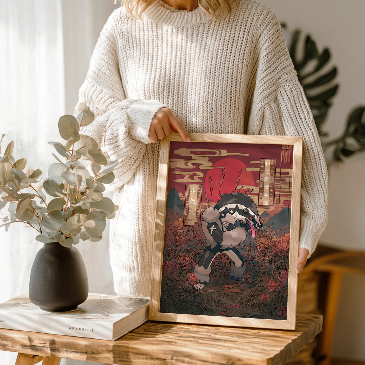 Person holding a framed artwork of Obstagoon Pokemon on a wooden table with a vase and book in the background.