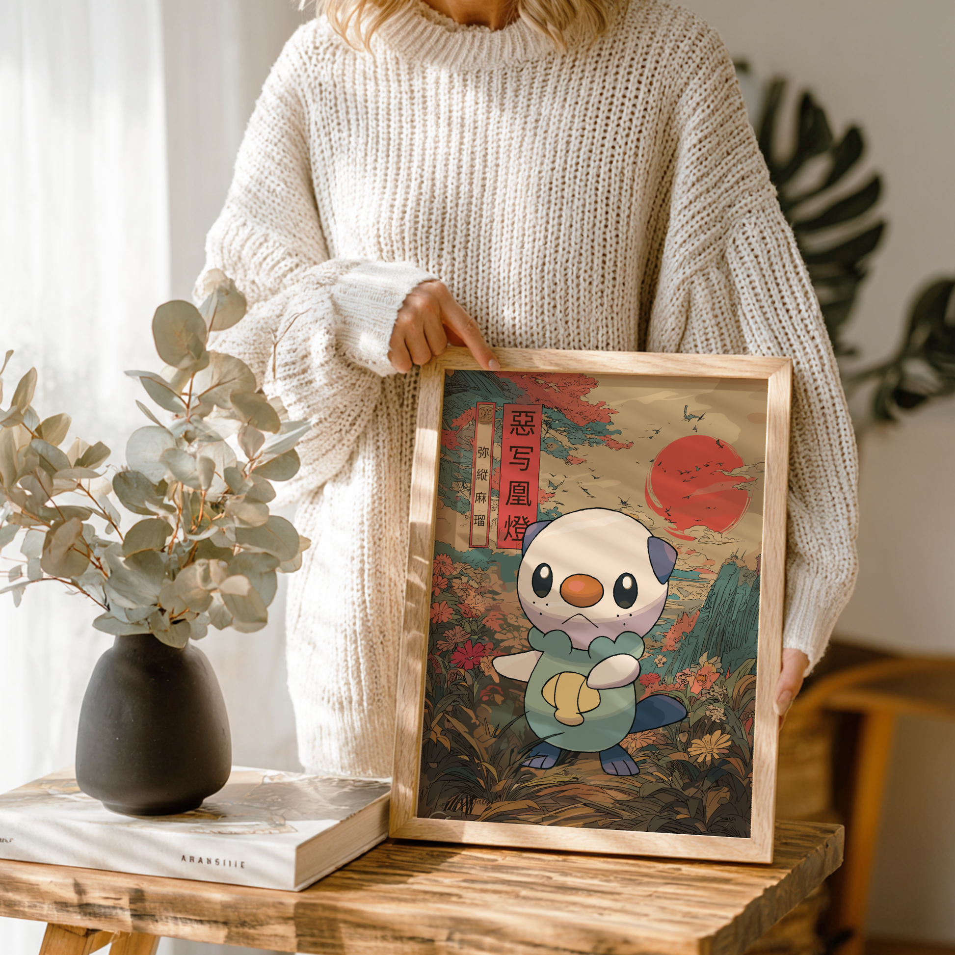 Person holding a framed illustration of Oshawott Pokemon character on a wooden table with a vase and book in the background.